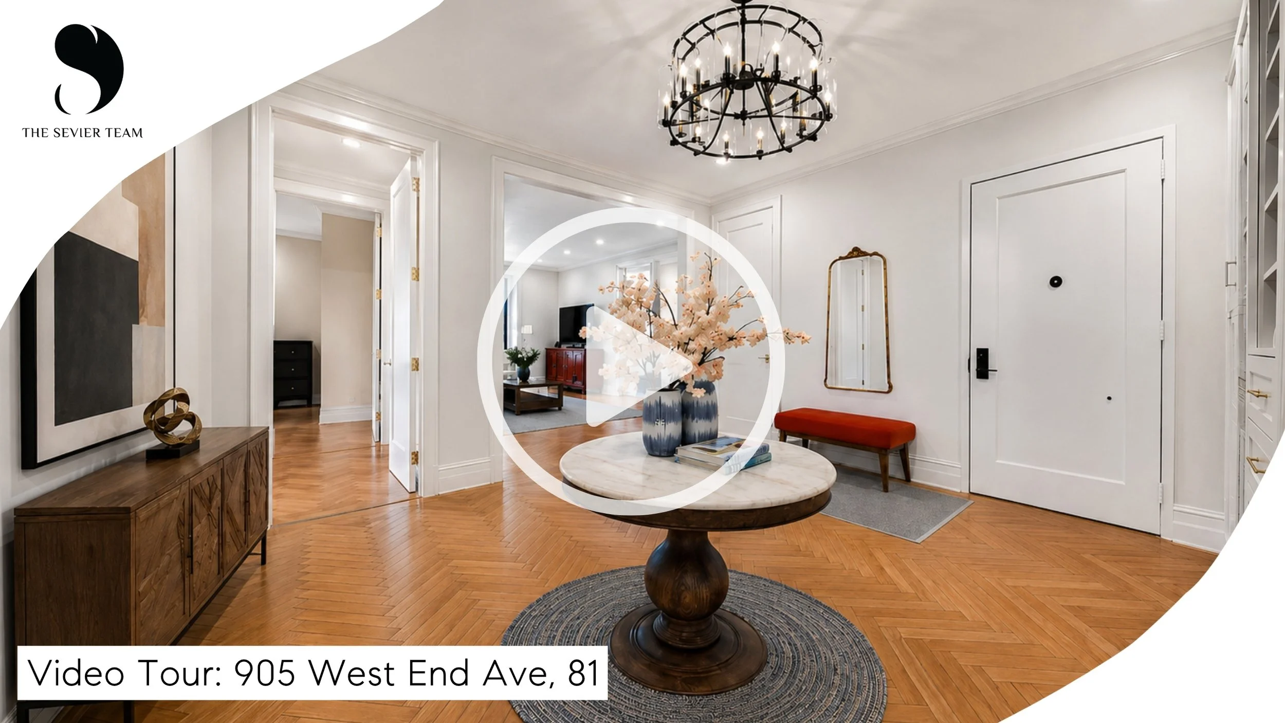 Interior view of a modern home with white walls, hardwood floors, a round marble-top table with flower vases, a red bench, and a chandelier, leading to a living room with a TV and decor.