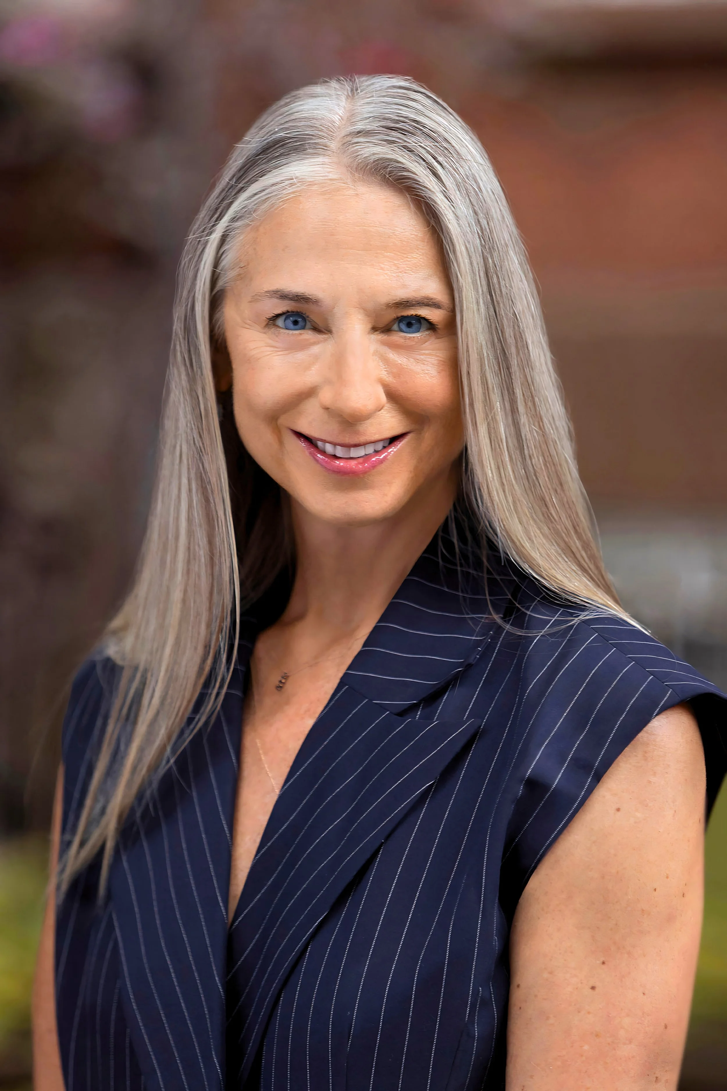 Close-up photo of a smiling woman with long, straight gray hair, blue eyes, and wearing a sleeveless navy blue blazer with thin pinstripes. Background is blurred outdoors.