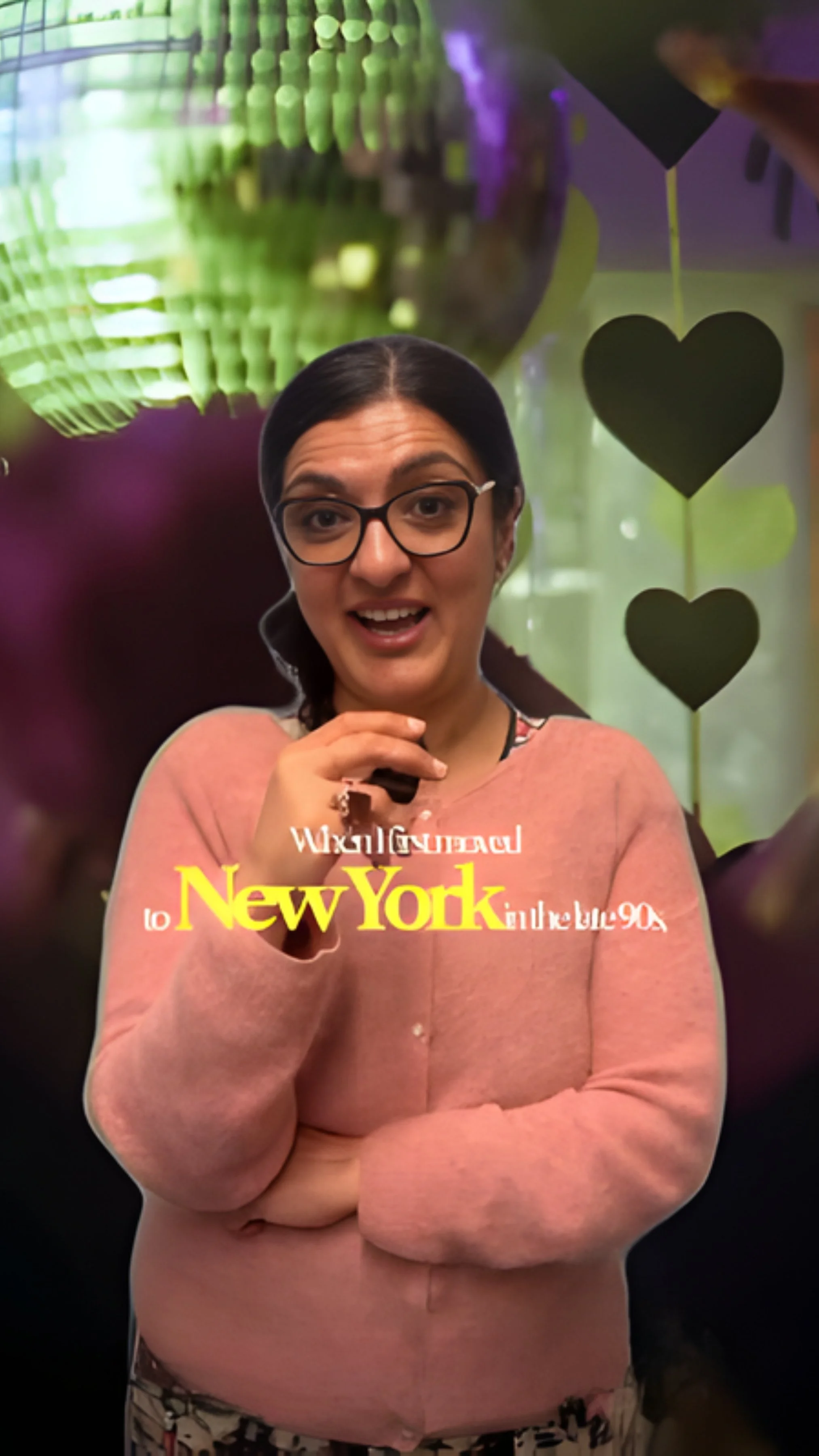 A woman with glasses and dark hair in a braid, wearing a pink sweater and floral pants, is smiling with her hand near her face. The background has colorful decorations including green and purple lights and hanging heart-shaped cutouts. The text overlay reads "New York."