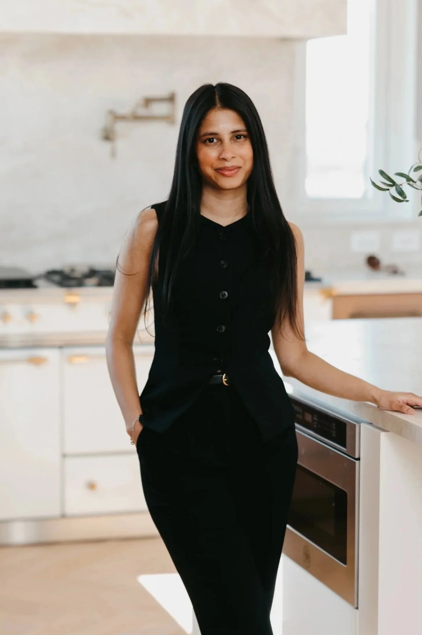 A woman with long black hair, wearing a sleeveless black button-up top and black pants, standing in a modern kitchen with white cabinets and a built-in oven, illuminated by natural light through a window on the right.