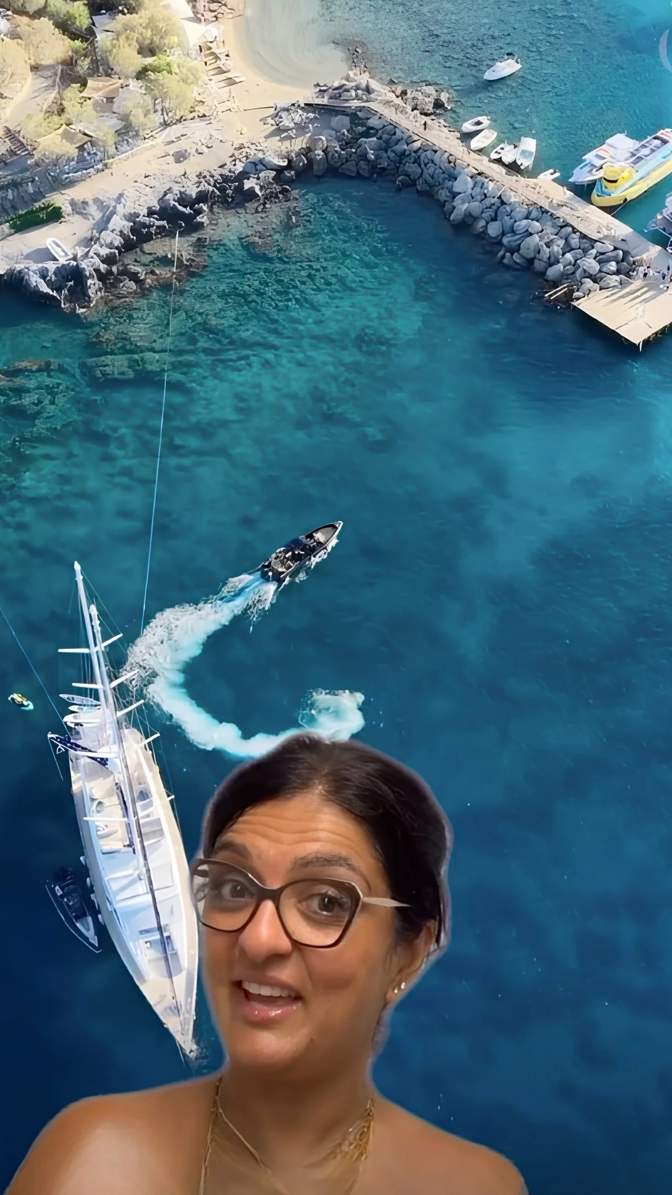 A woman taking a selfie at a marina, with a large yacht, boats, and a rocky coastline in the background.