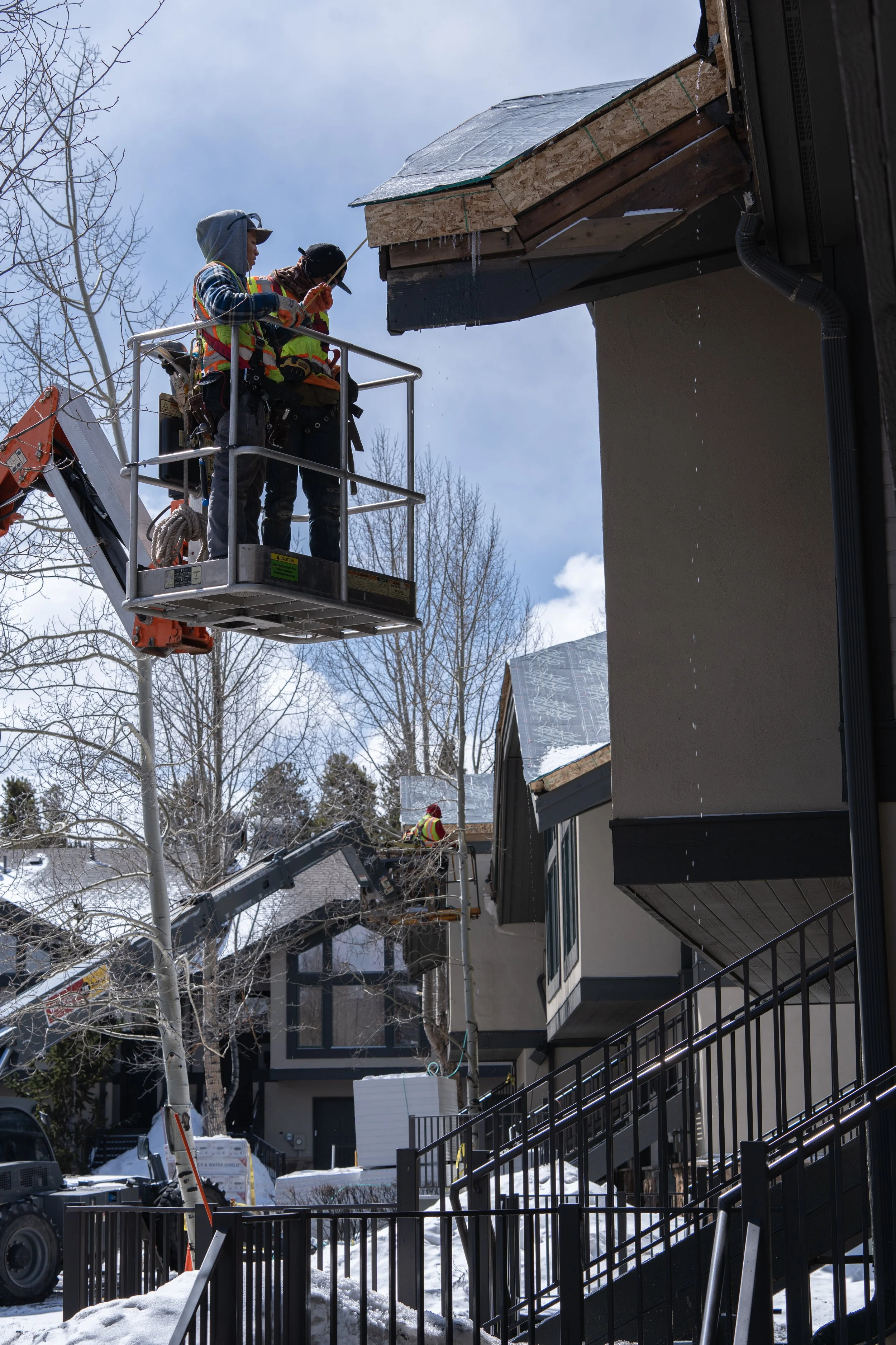 Construction workers in high-visibility clothing on a hydraulic lift, working on a roof in a snowy residential area.