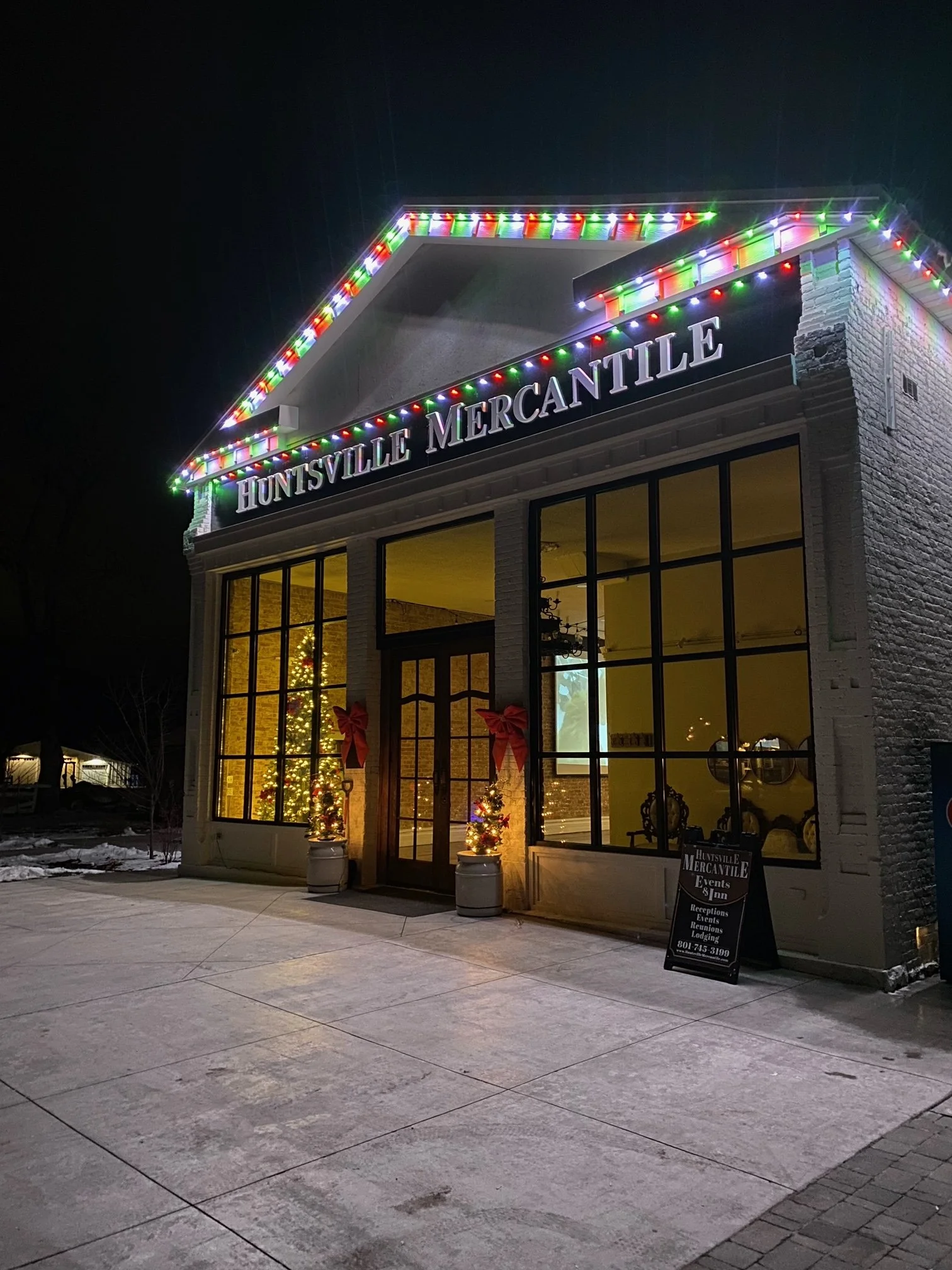 Night view of Huntville Mercantile storefront decorated with Christmas lights and holiday wreaths, with a Christmas tree visible inside