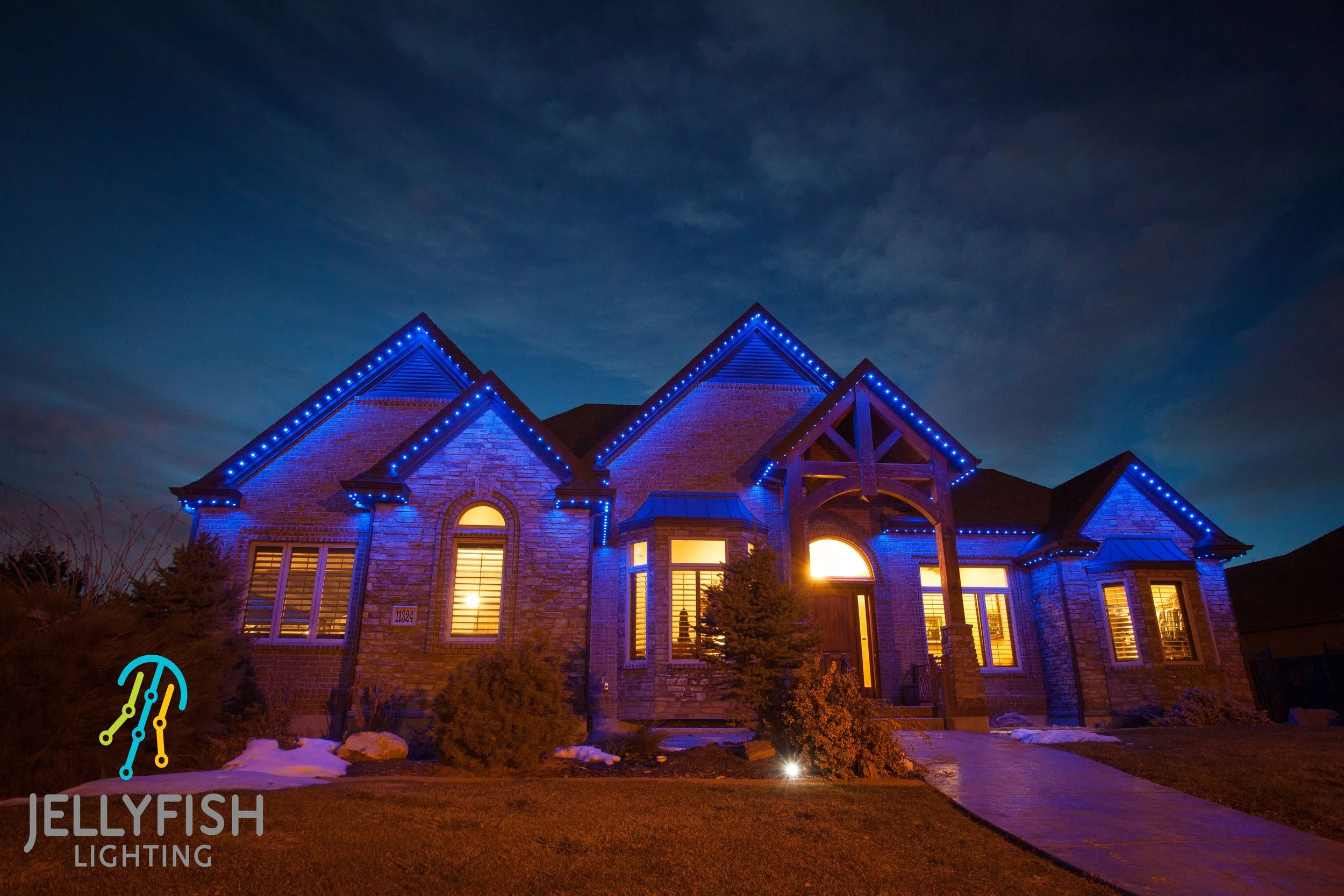 A house at night is decorated with blue string lights along the rooflines and windows, with warm yellow interior lights visible through the windows and a lit pathway leading to the front door.