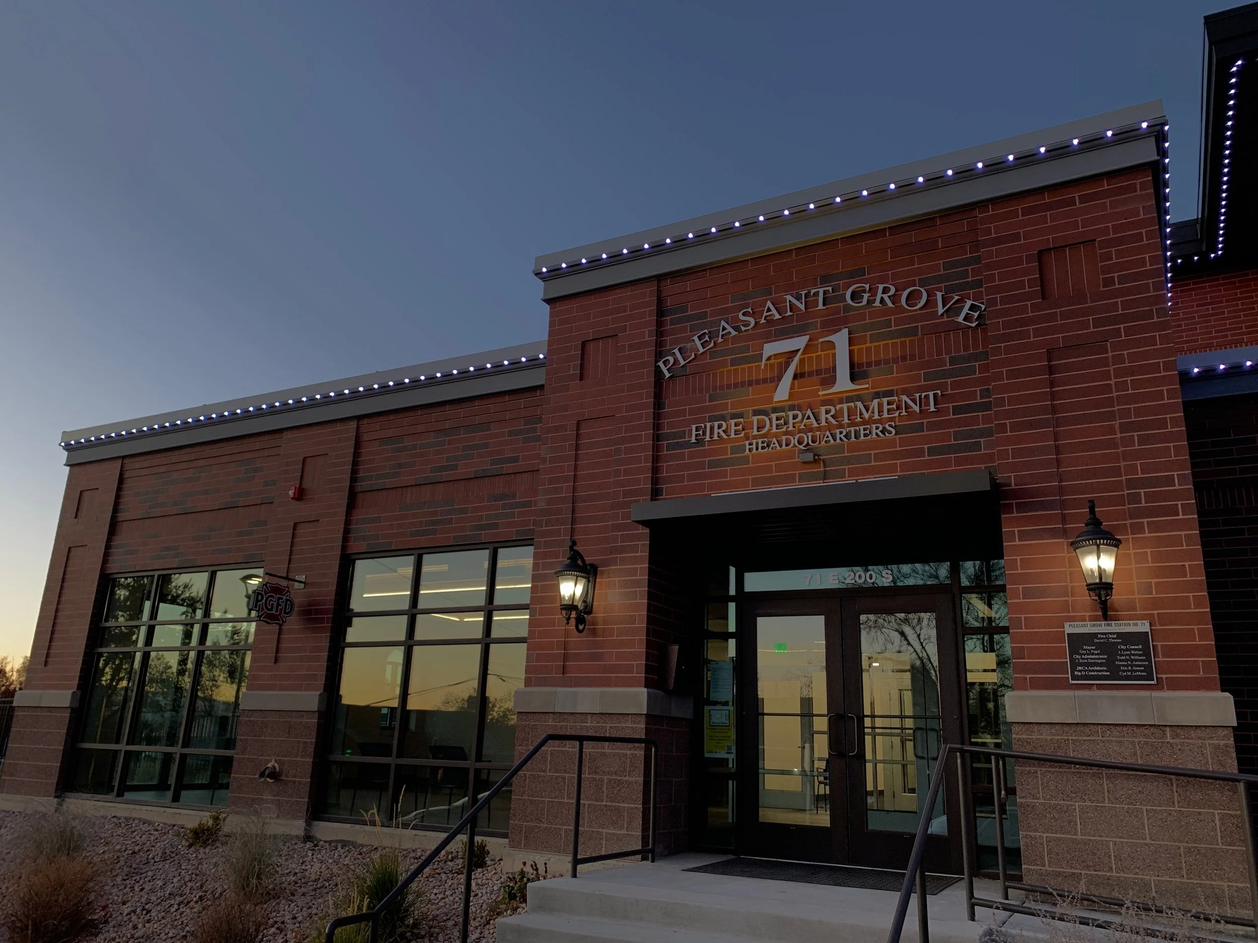 Brick building with sign reading 'Pleasant Grove 71 Fire Department Headquarters'; exterior lit with string lights and wall-mounted lanterns; large glass entrance doors; evening setting with clear sky.