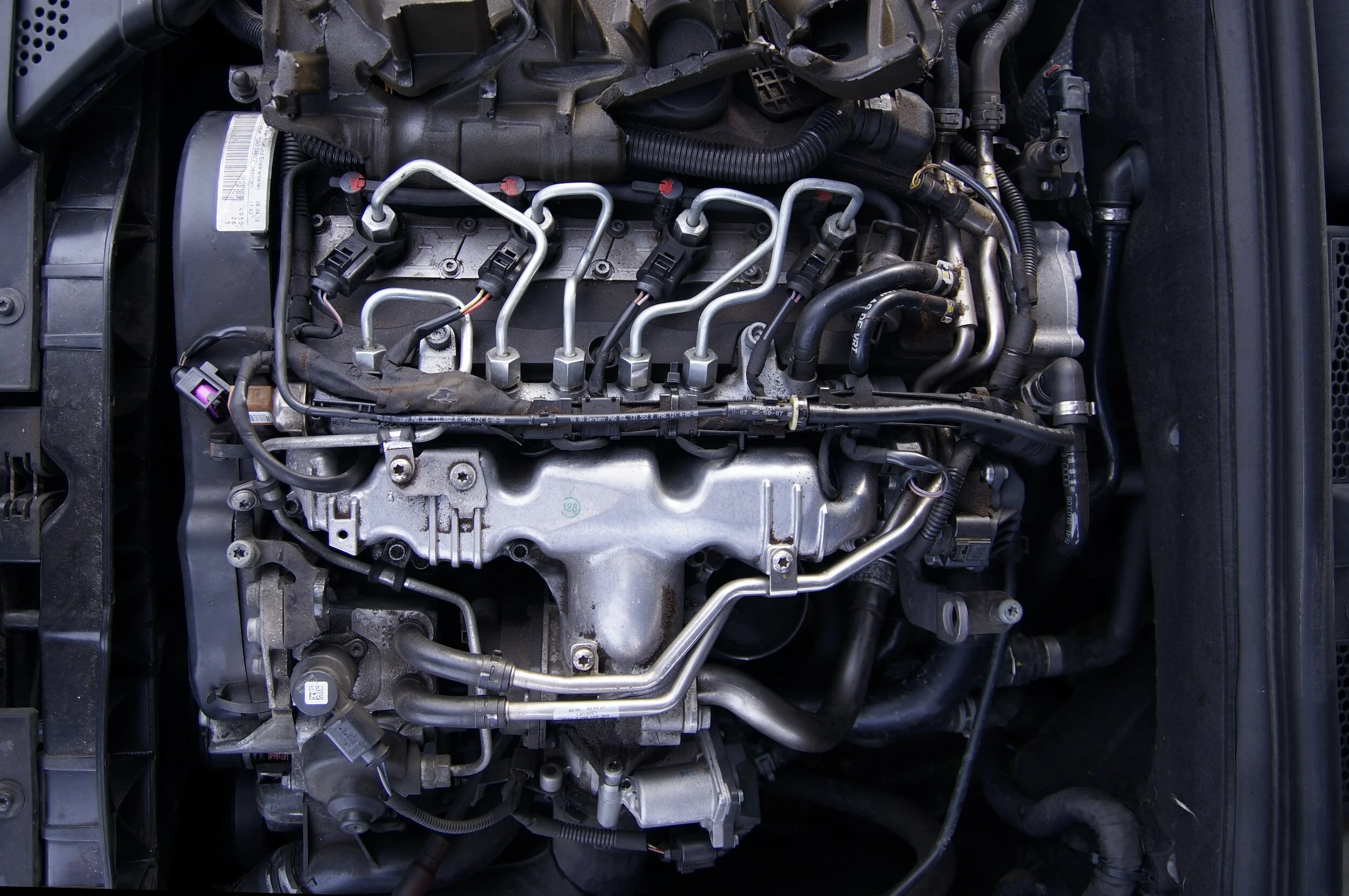 A car engine with various metal and rubber components, hoses, and wiring visible.