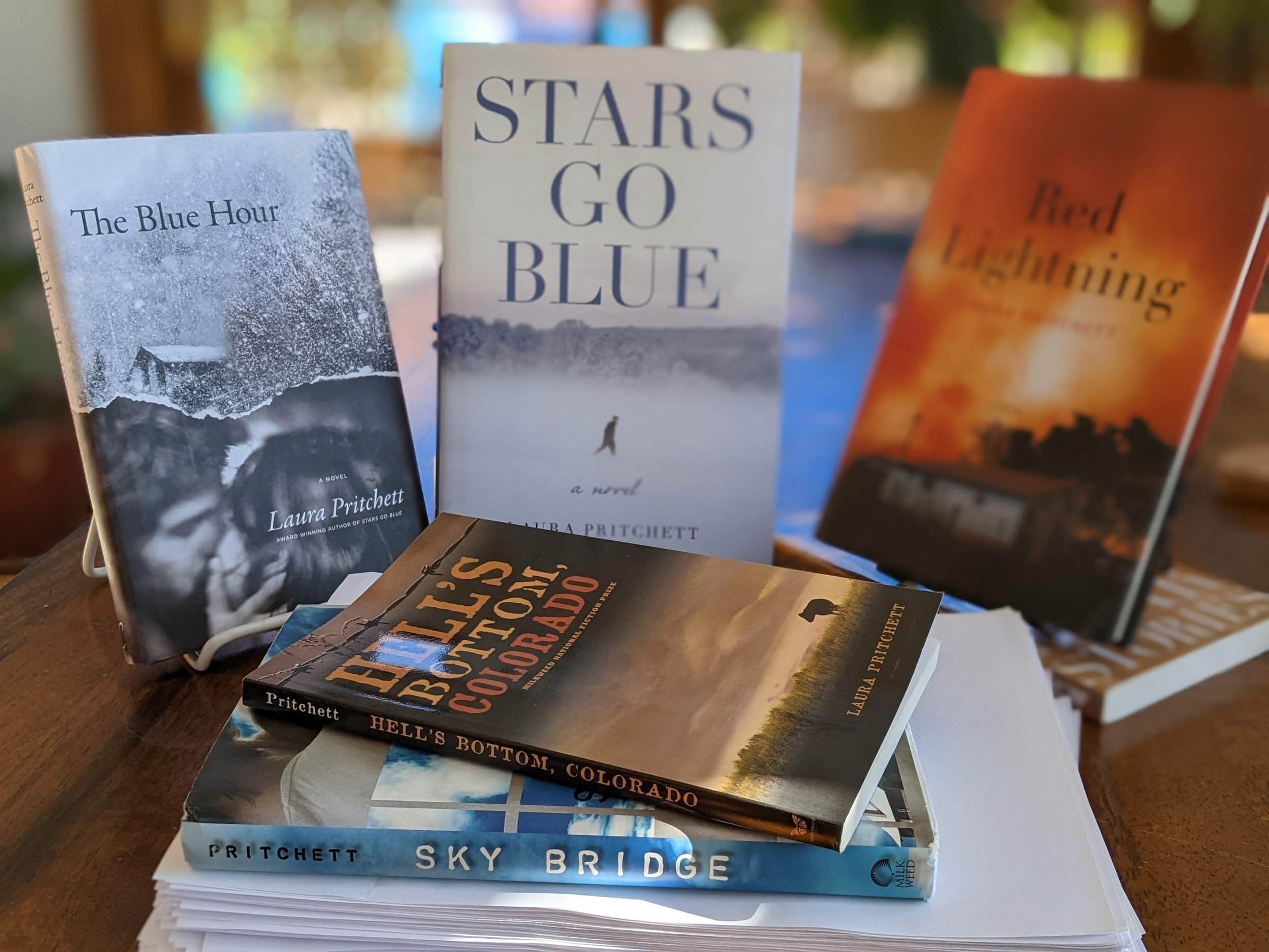 Several of Laura's books sit on a table. From left to right: The Blue Hour, Stars Go Blue, Red Lightning, Hell's Bottom Colorado, Sky Bridge.