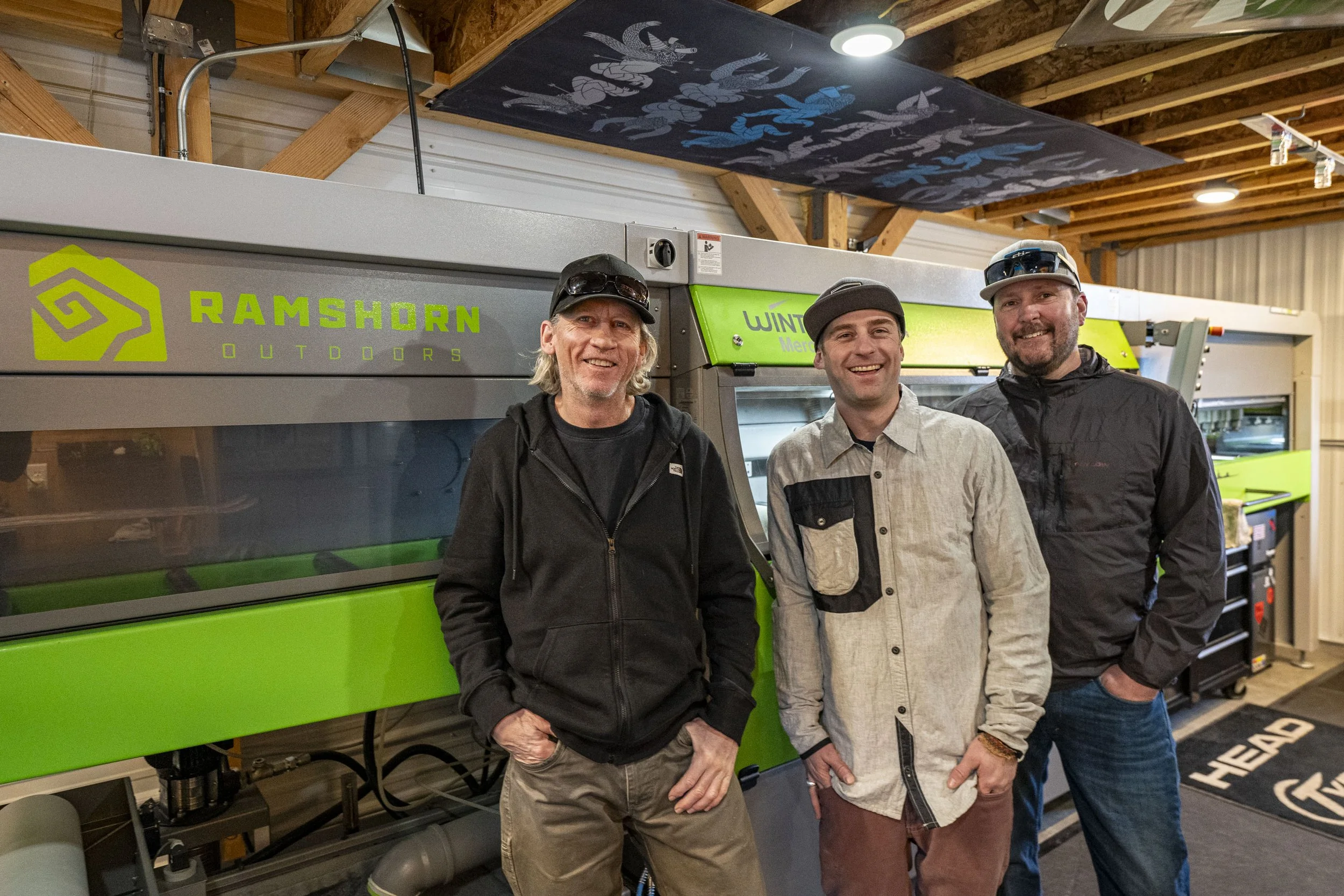 Three men standing in front of green and gray Ramshorn Outdoors equipment in a wooden building, smiling at the camera.