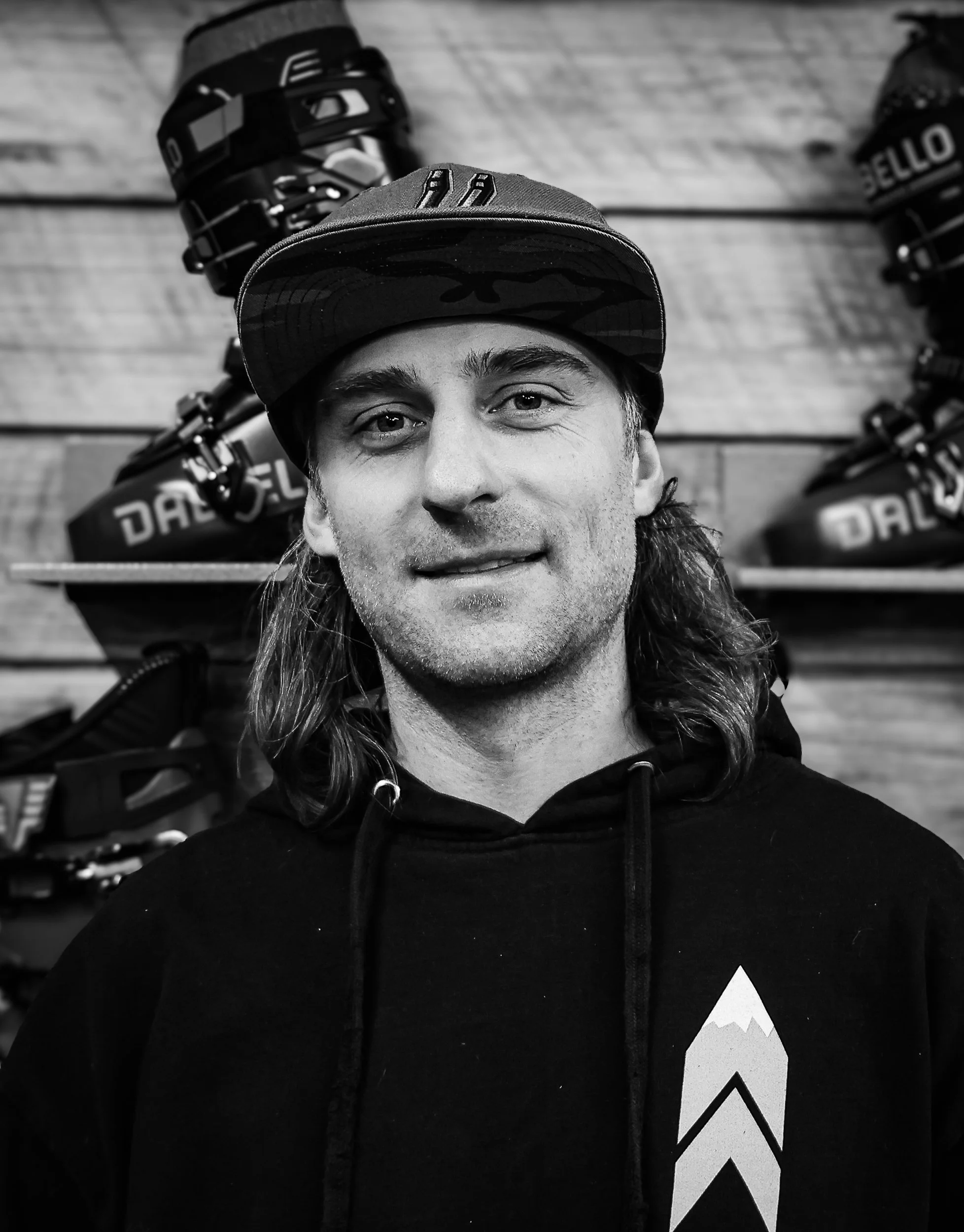 A man with long hair and a cap, smiling, standing in front of sporting equipment on a wooden wall.