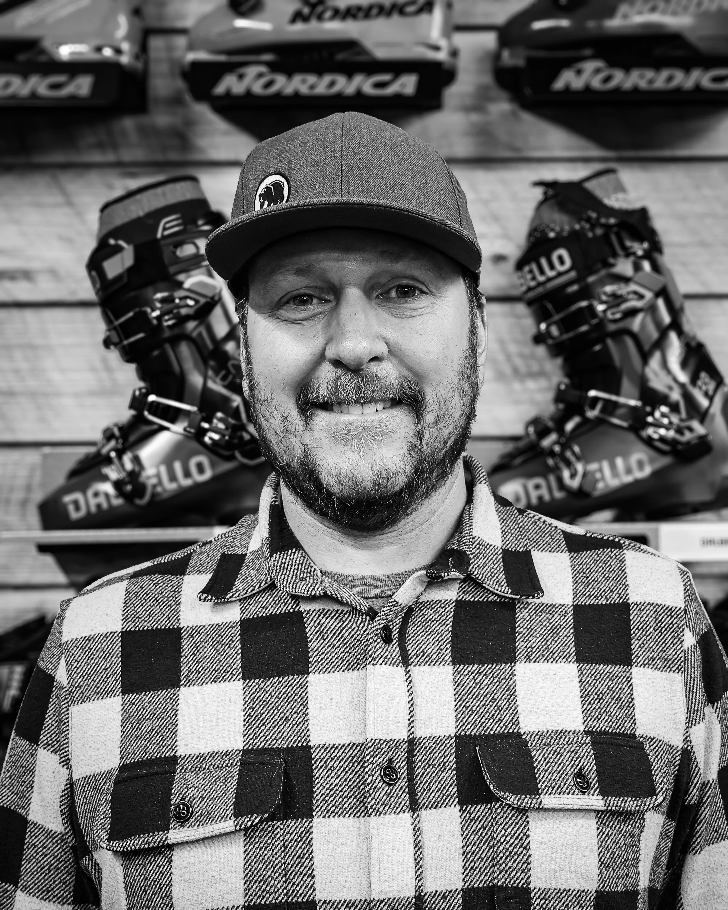 A man with a beard smiling, wearing a checkered shirt and a baseball cap, standing in front of a wood-paneled wall with ski boots and snowboard boots displayed behind him.