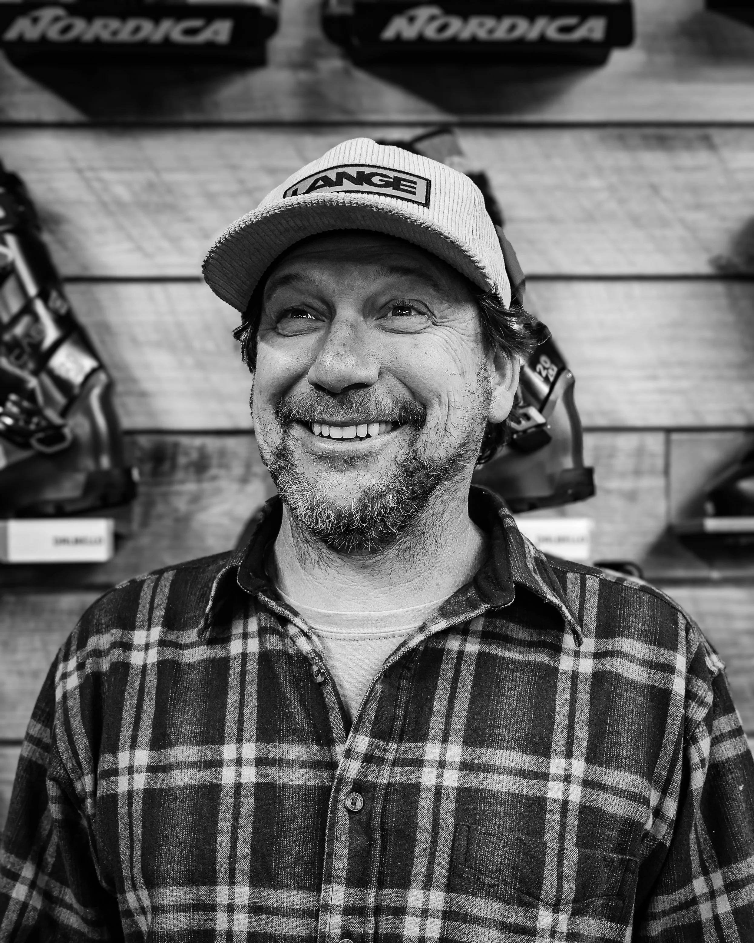 A smiling man with a beard wearing a baseball cap and checkered shirt, standing in front of a display of snowboarding or skiing equipment.