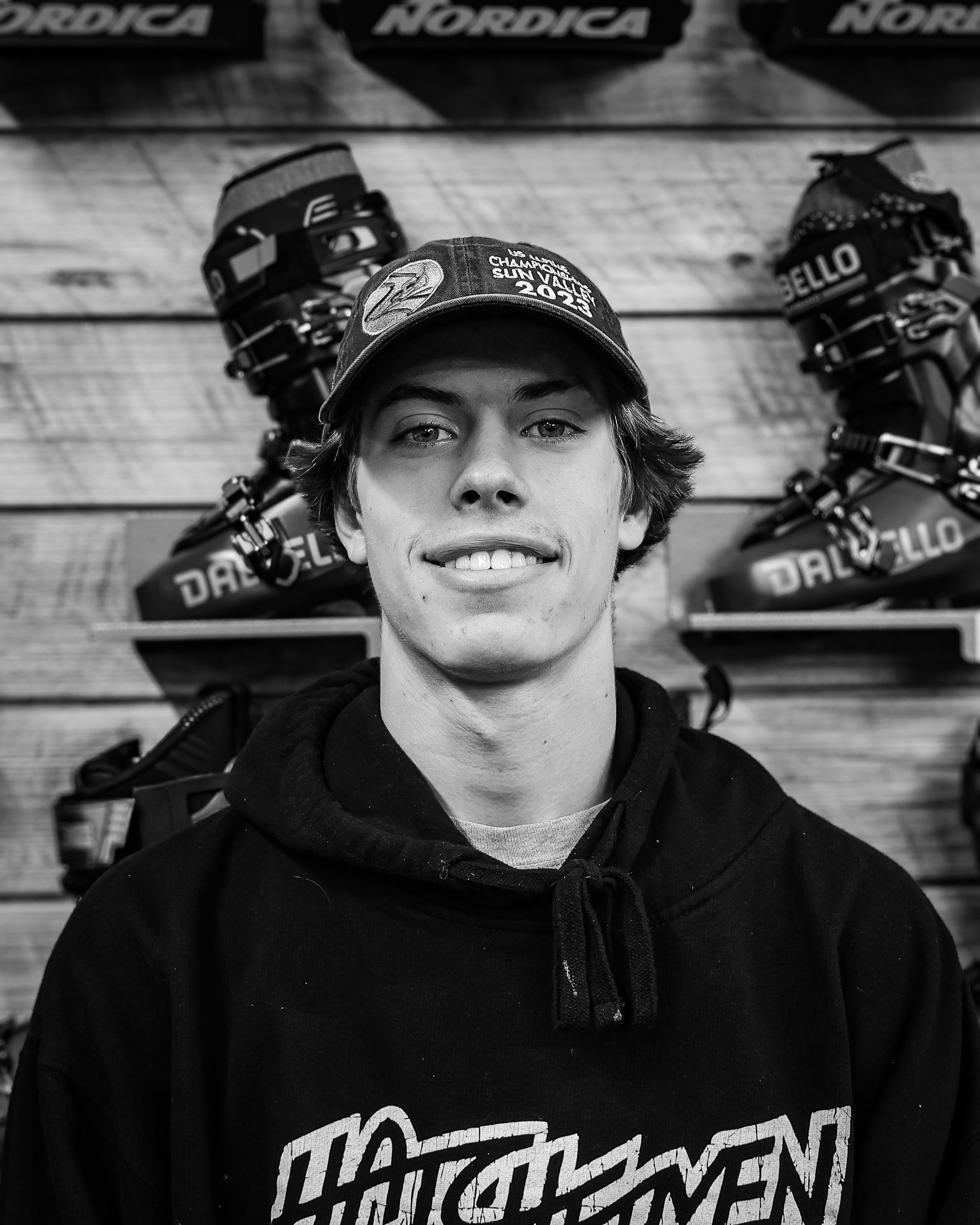 A young man with wavy hair wearing a cap and hoodie, smiling, standing in front of a wall with ski boots displayed on shelves, in black and white.