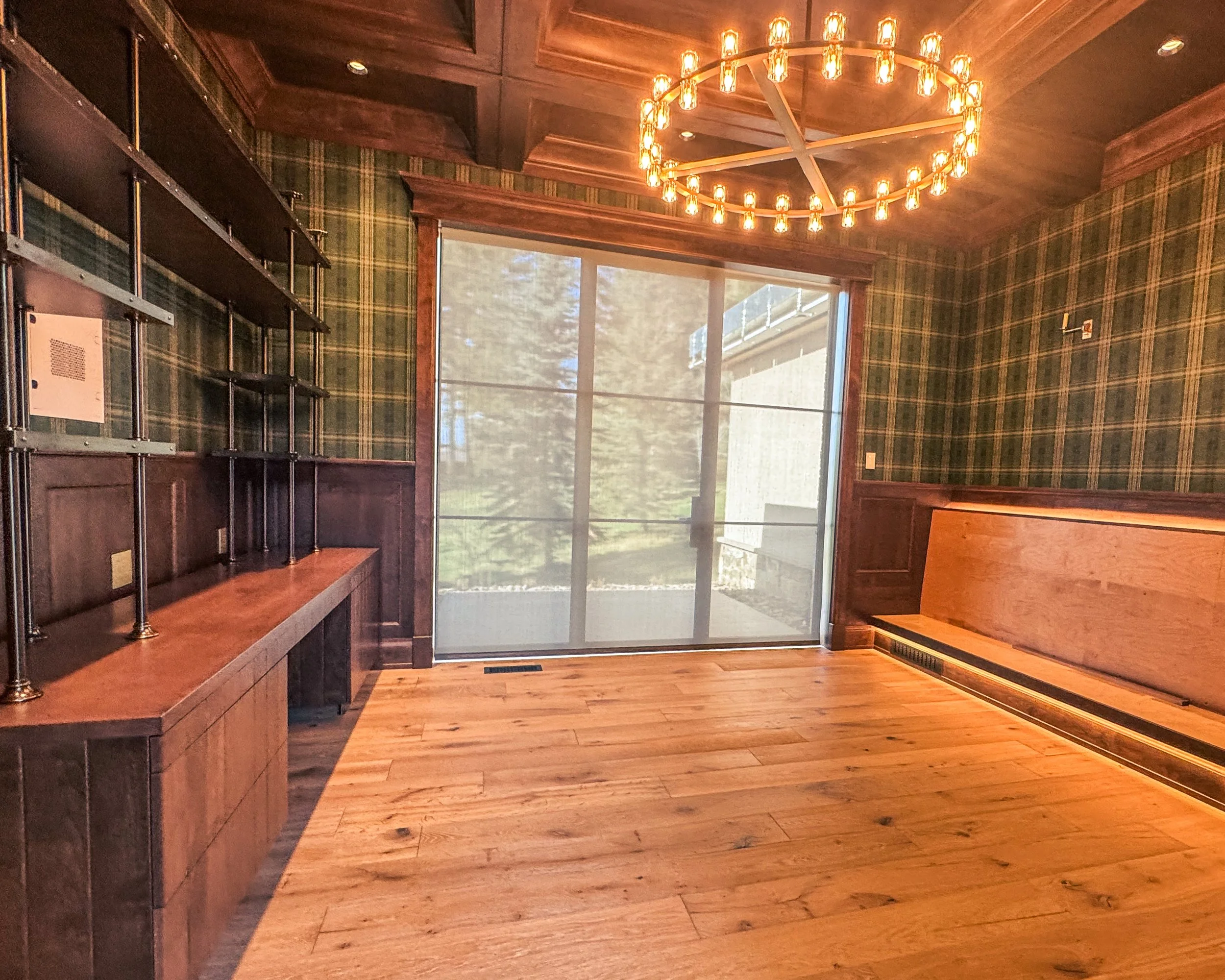 Empty room with wood-paneled walls, patterned green plaid wallpaper, a large window with a screen, a wooden bench, and a circular chandelier with multiple light bulbs.