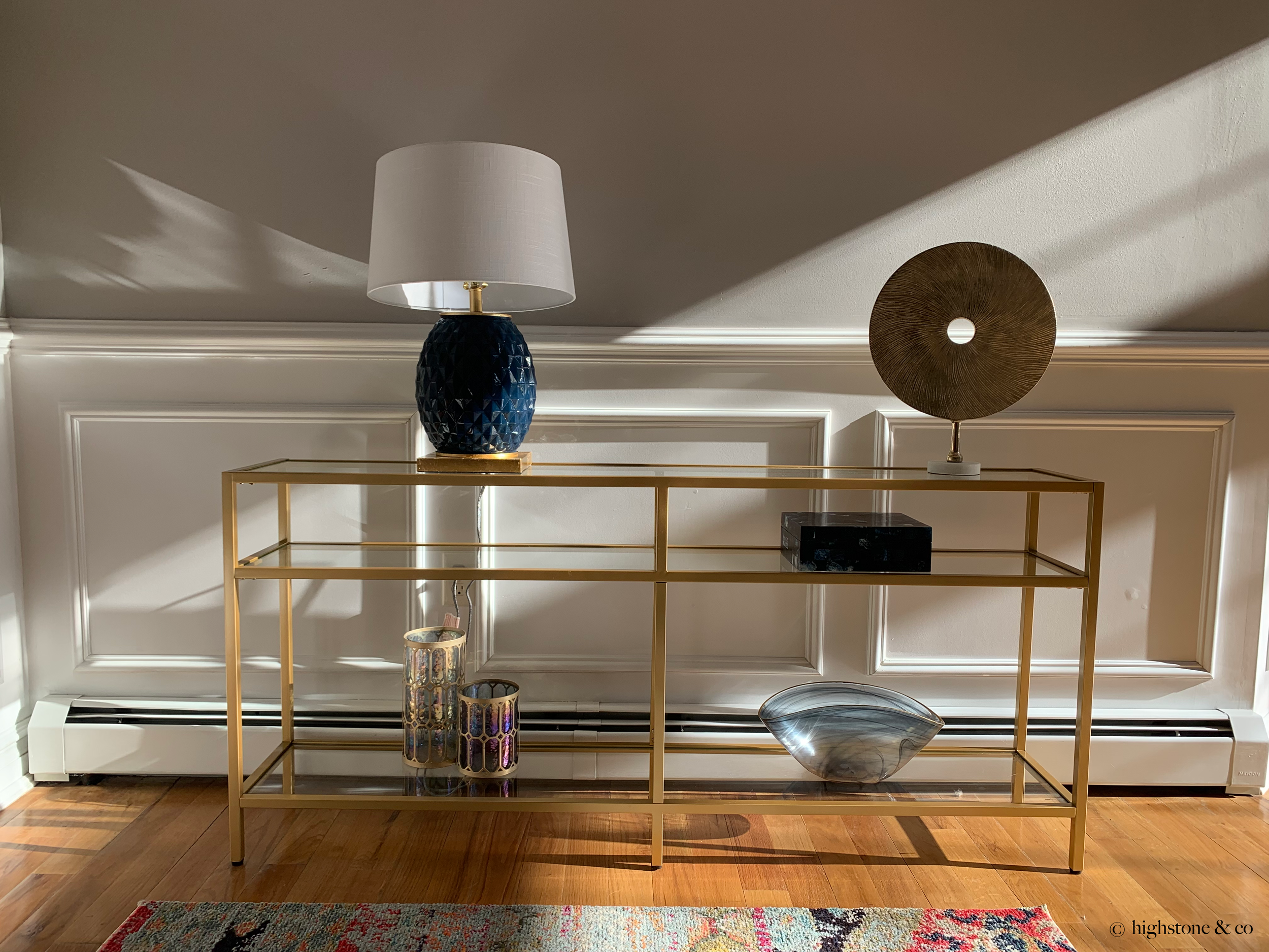 A gold metal side table with a glass top holding a blue ceramic lamp with a white lampshade and a decorative gold disc sculpture. On the lower shelf are two glass candle holders with a honeycomb pattern and a metallic finish, along with a large gray and white shell. The table is set against a white wall with molding, with sunlight casting shadows.