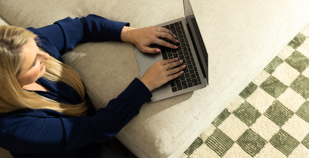 A woman with long blonde hair working on a laptop while sitting on a beige couch.