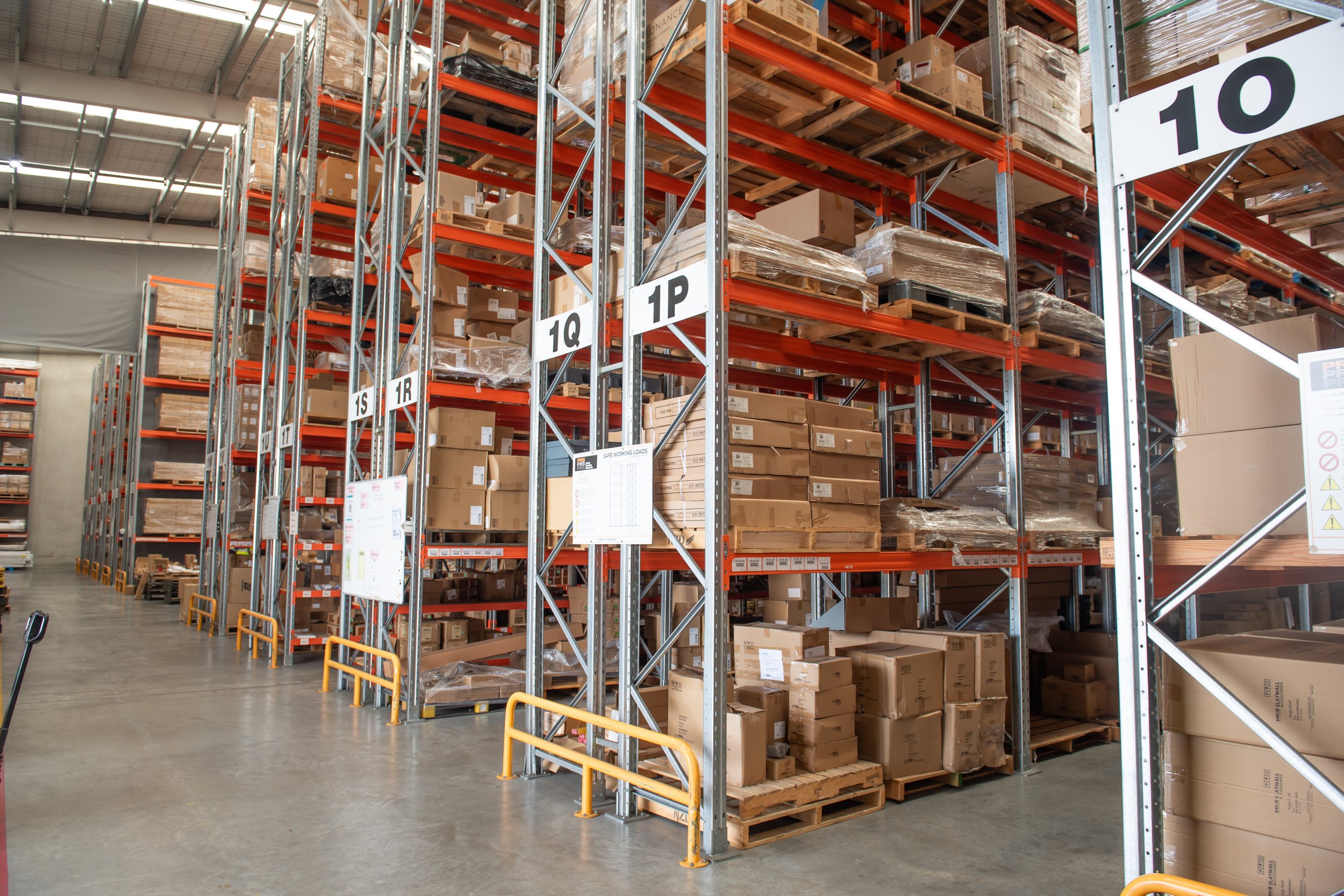 Warehouse aisle with tall orange and gray metal shelves filled with numerous cardboard boxes, some wrapped, arranged in various levels. Signage indicates aisle numbers.