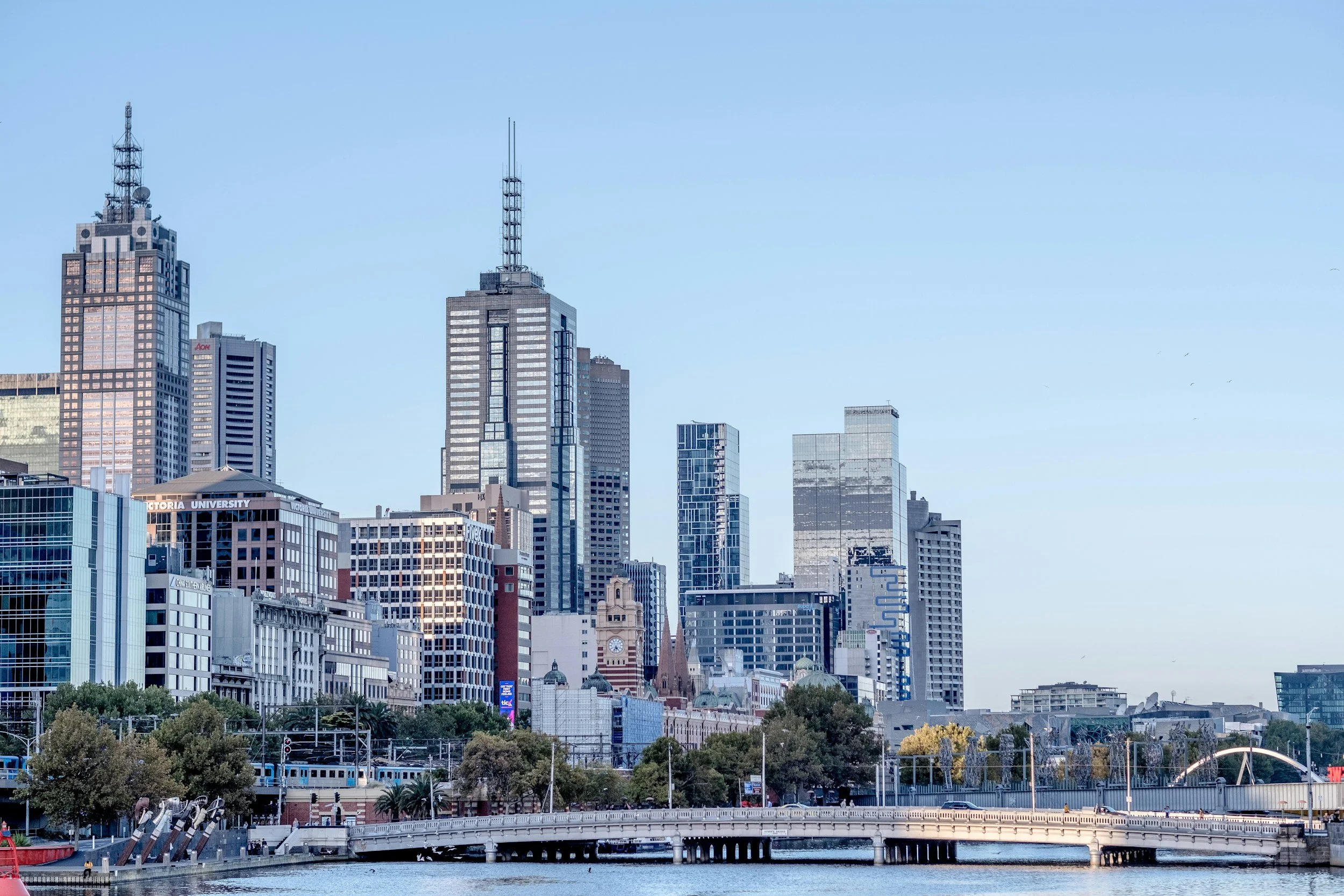 City skyline with tall skyscrapers, including the Eureka Tower, near a river with a bridge and trees in the foreground.