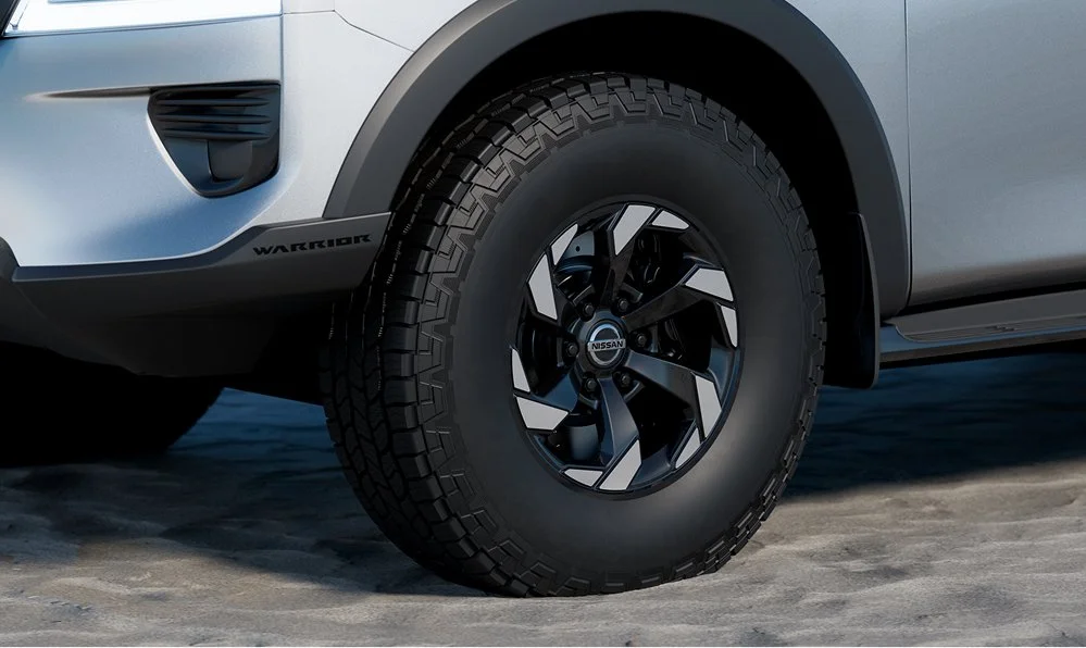 Close-up of the front wheel of a Nissan SUV, featuring a black and silver alloy rim and large all-terrain tire. Part of the vehicle's front bumper and side panel are visible.