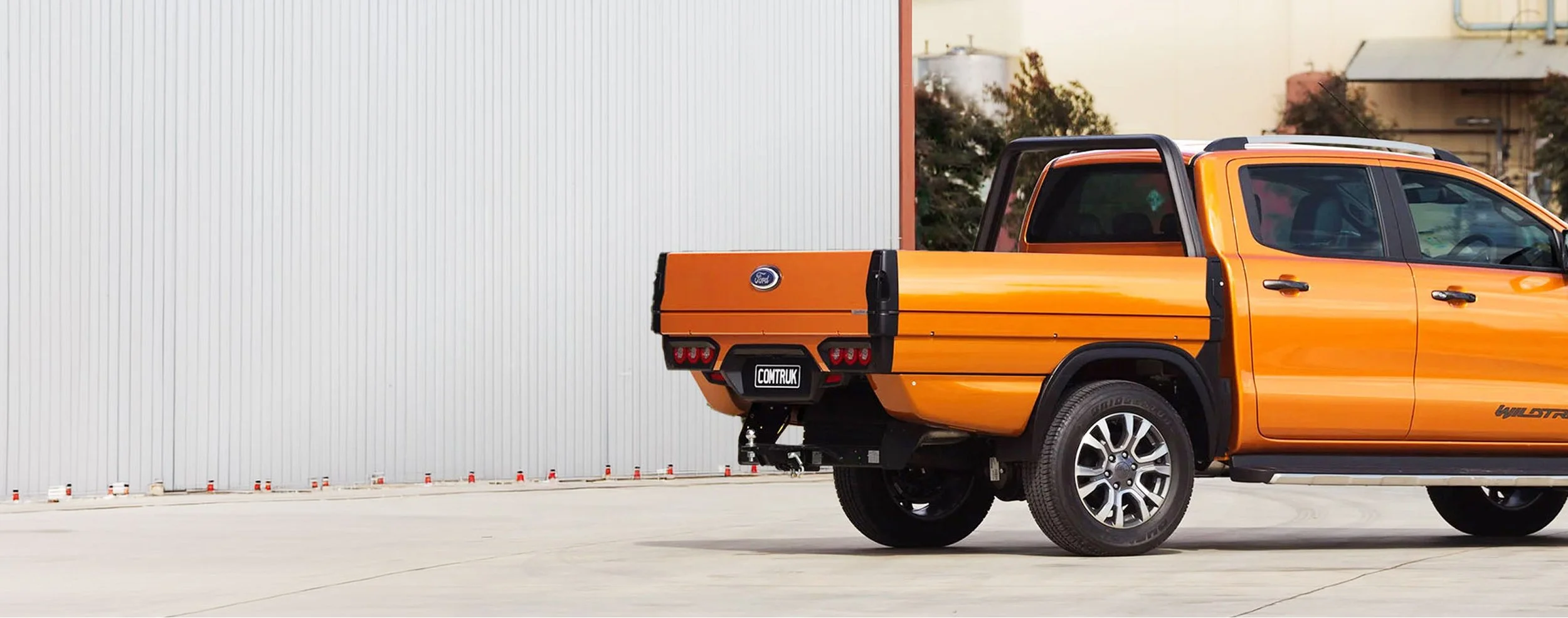 Orange Ford pickup truck with black accents parked on concrete in front of a large white industrial building with a red trim and some trees in the background.