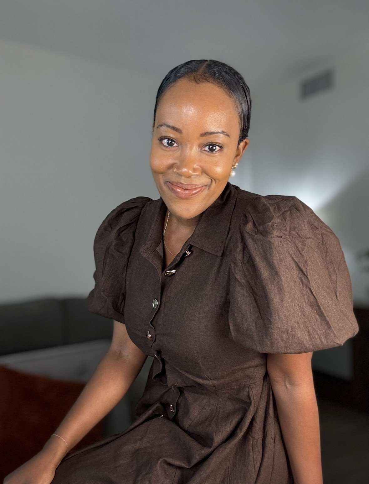 A smiling woman with dark hair styled close to the scalp, wearing a dark brown dress with puffed sleeves, sitting in a room with a couch and a plant in the background.