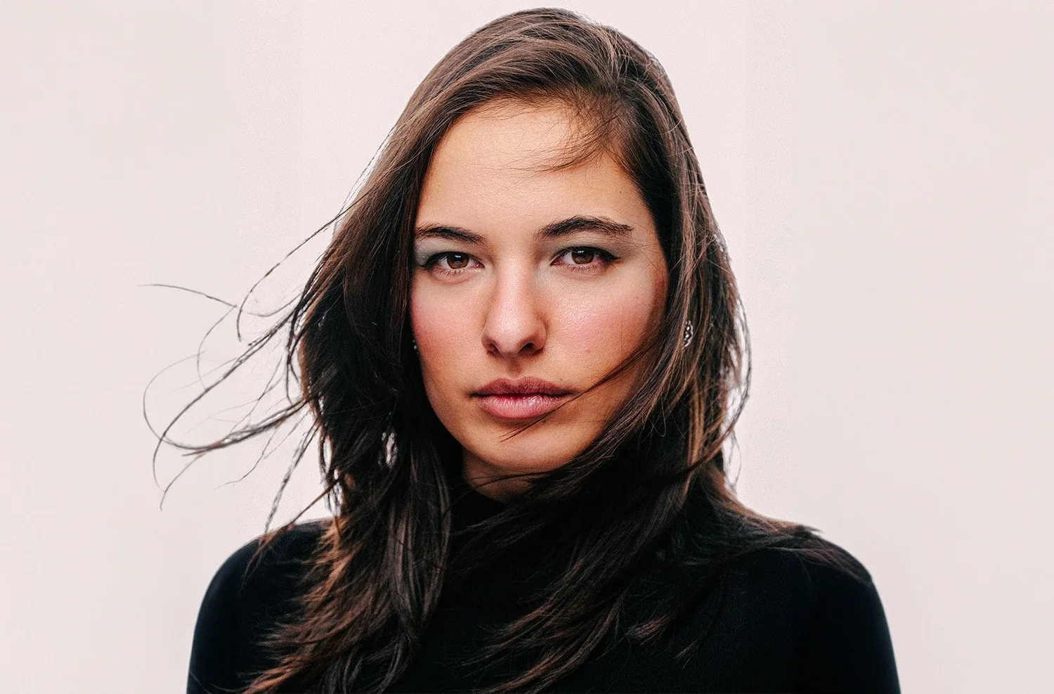 A woman with long brown hair, wearing a black top, looking directly at the camera with a neutral expression.