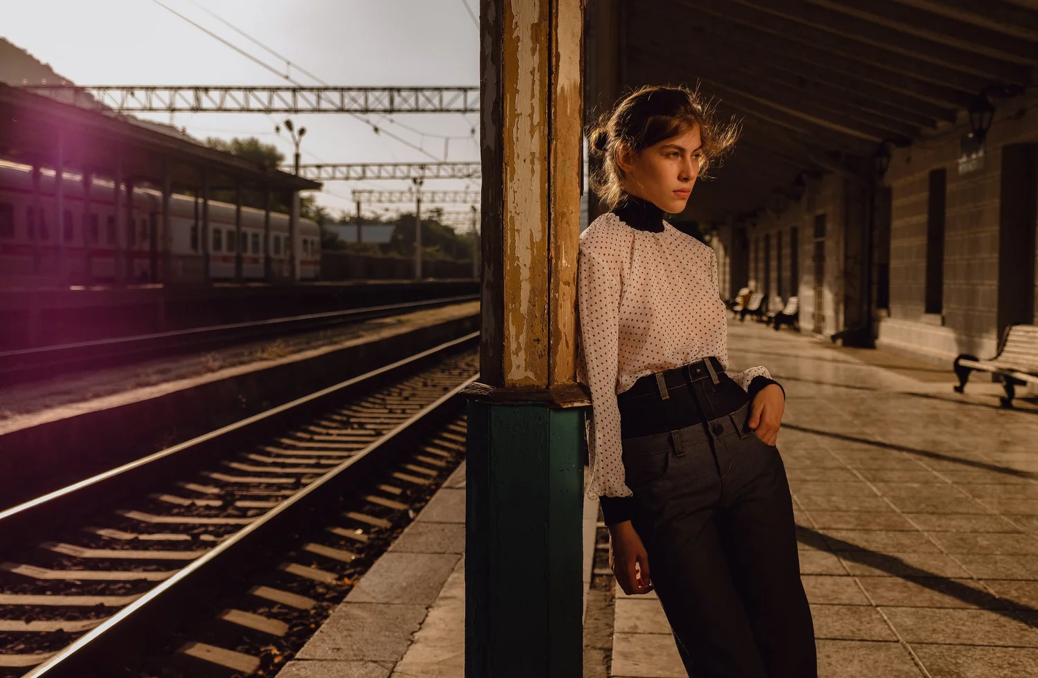 A young woman standing at a train station platform, leaning against a weathered wooden post, with a train in the background, during sunset.