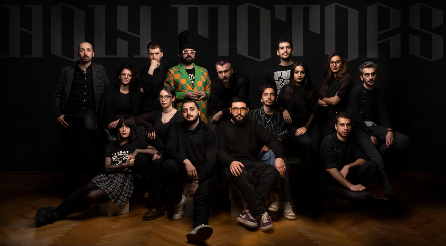 A group of fifteen diverse young adults posing together against a dark background with large, stylized lettering.