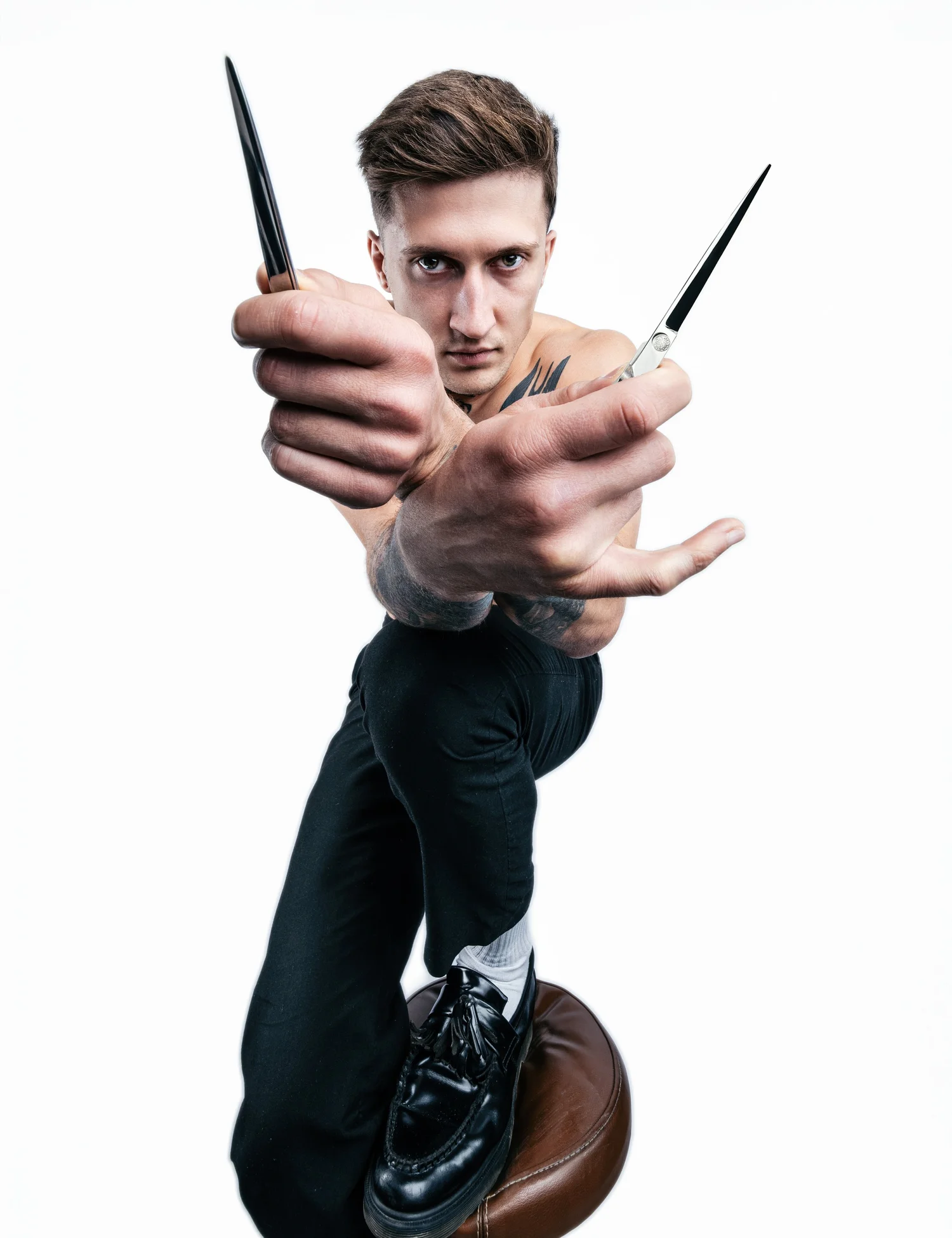 A man balancing on a round, brown stool, holding a pair of scissors and a straight razor, with an intense look, wearing black pants, black shoes, and white socks, against a plain white background.