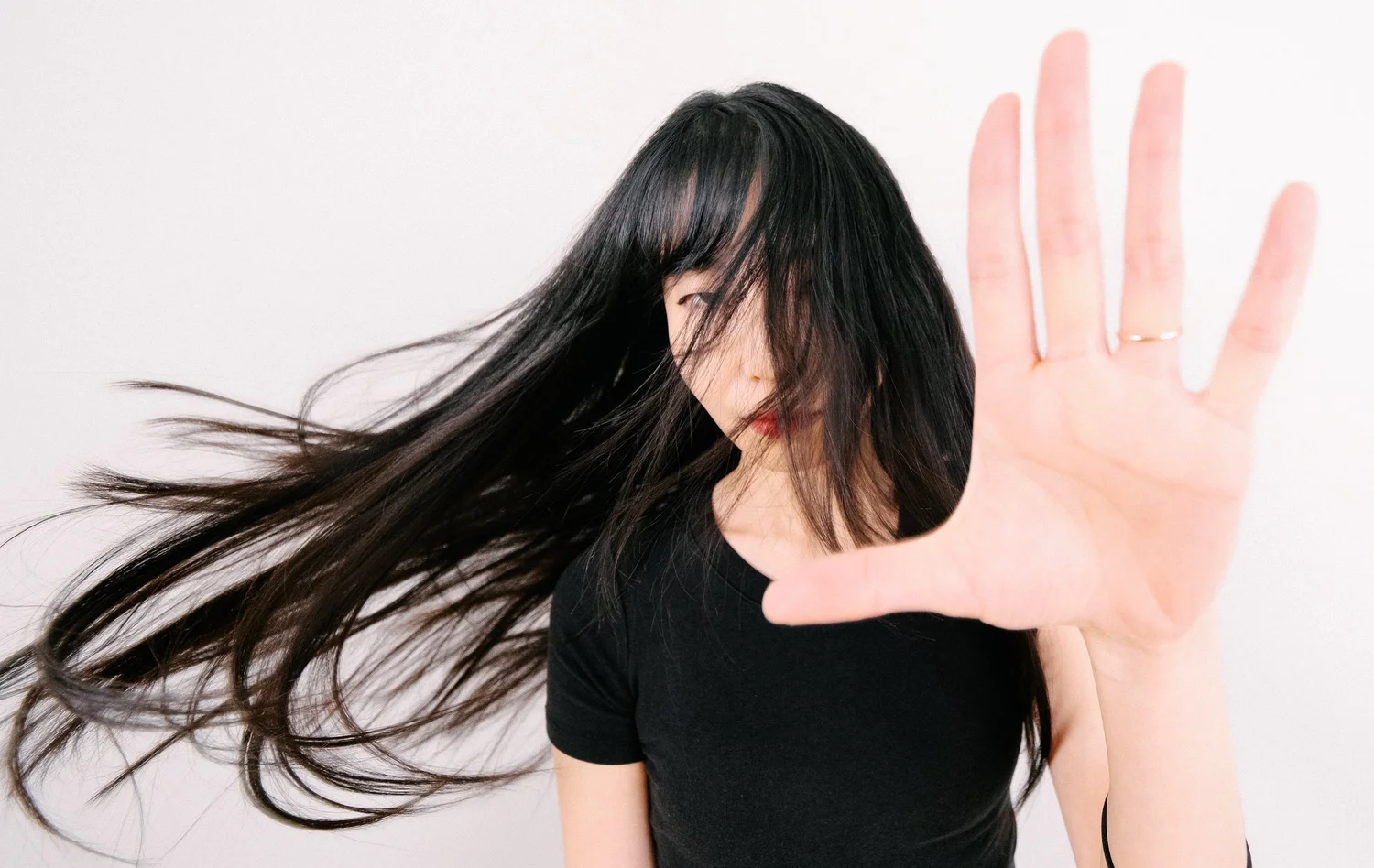 Young woman with long black hair covering part of her face, wearing a black shirt, holding her hand up towards the camera with fingers spread apart, against a white background.