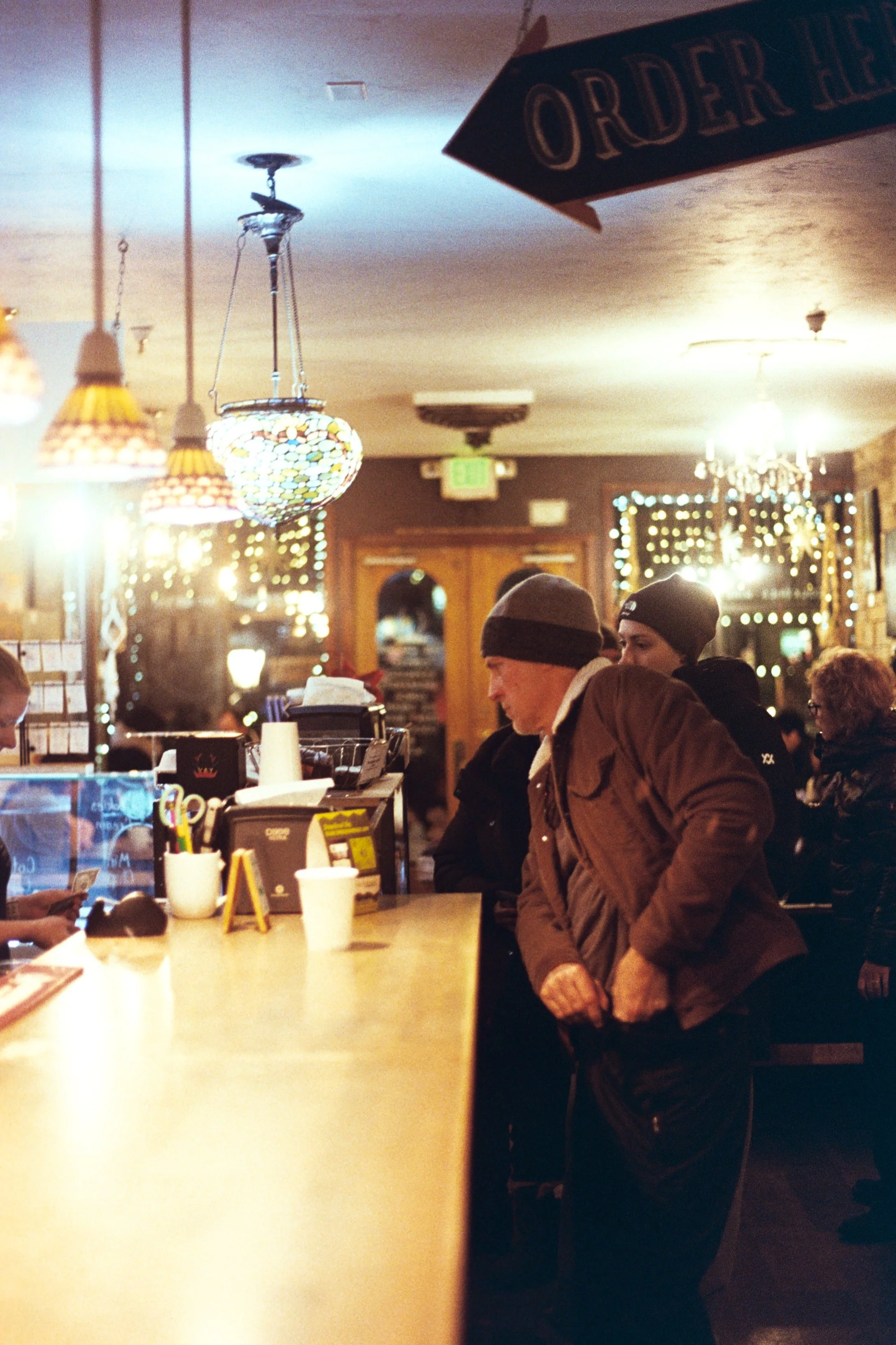 People standing at a restaurant counter with warm lighting and decorative hanging lamps, ordering food.