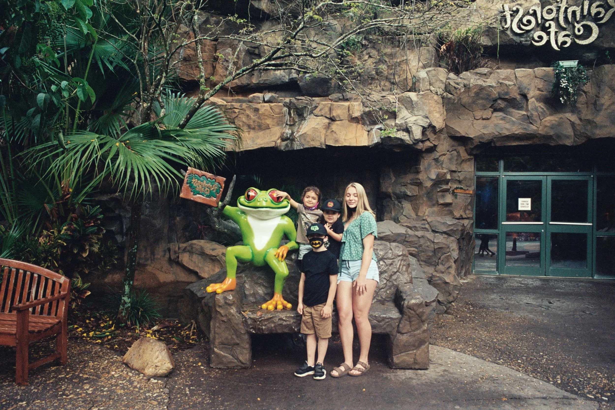 Three children and a young woman posing with a large cartoonish frog statue holding a sign. They are in front of a rocky, jungle-like backdrop.