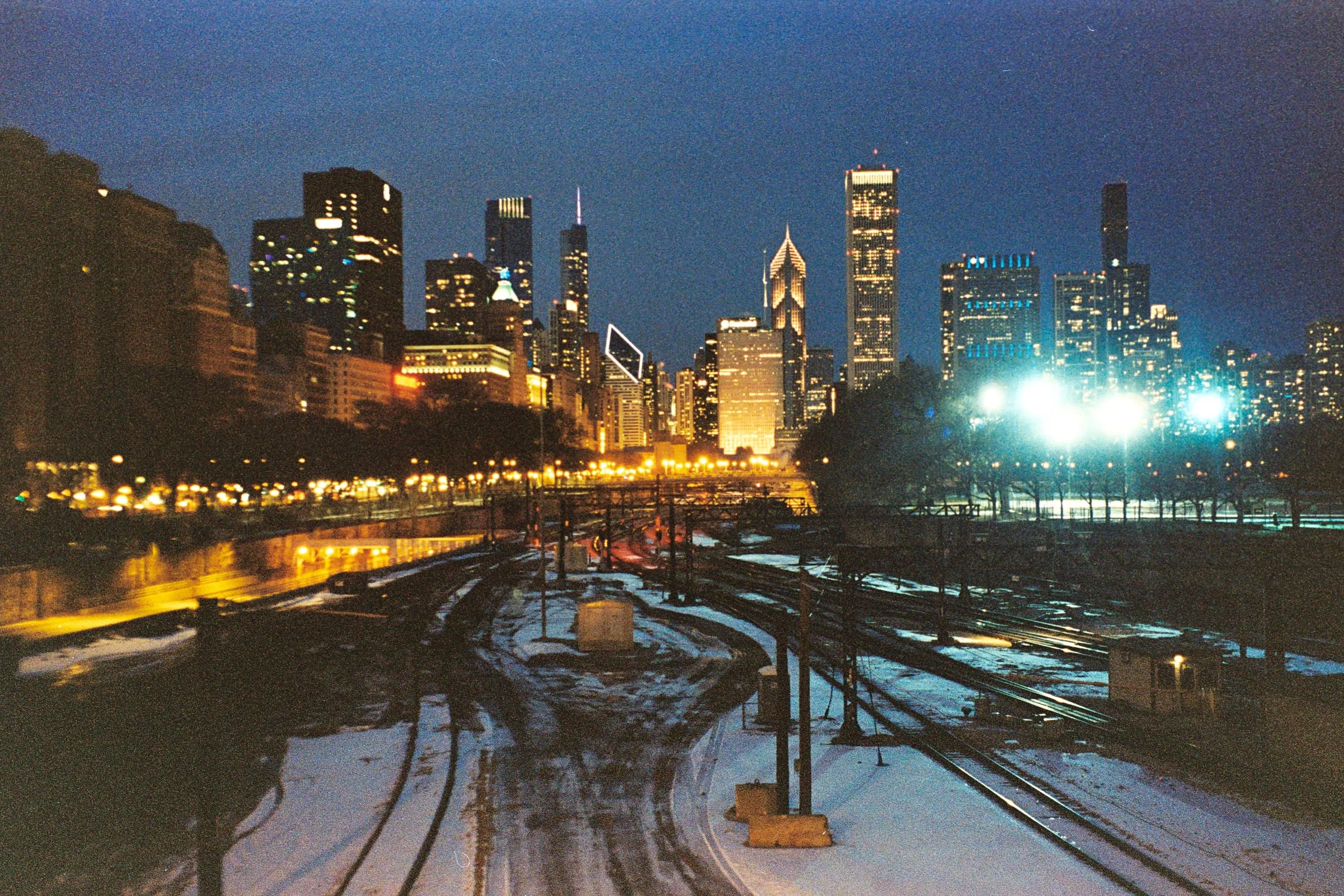 Nighttime cityscape of Chicago with illuminated skyscrapers, snow-covered train tracks in the foreground, and bright artificial lights.