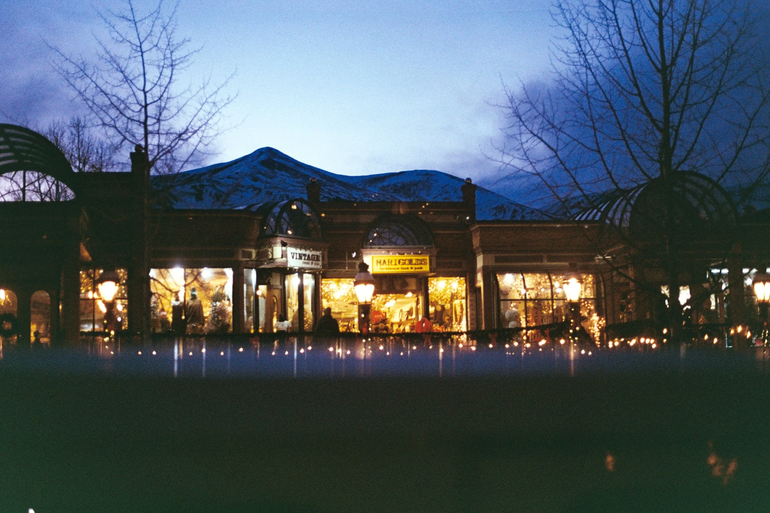 A shopping mall at dusk with snow-capped mountains in the background, trees without leaves, and shops with illuminated windows and string lights.