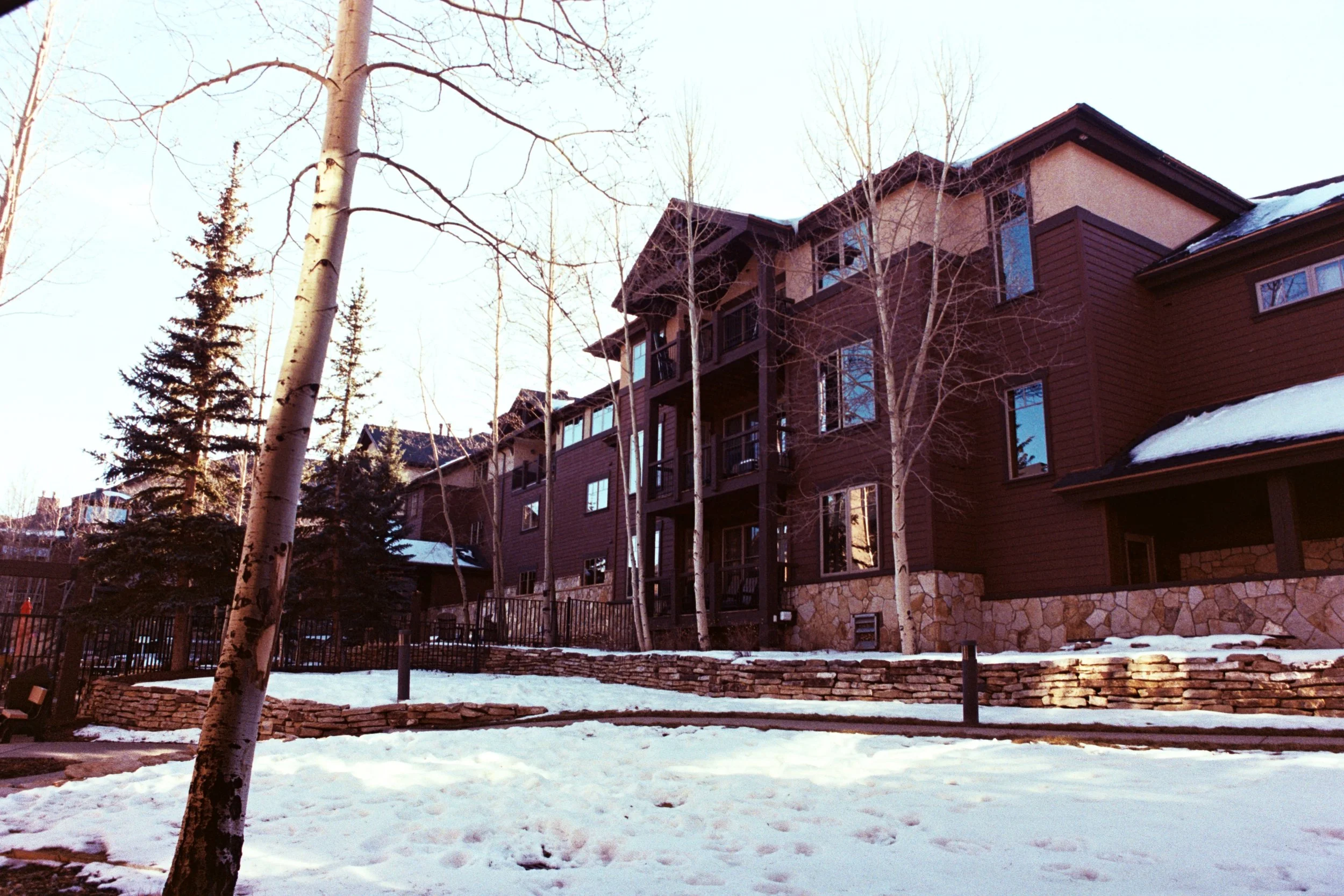 A multi-story residential building with wooden siding, stone foundation, and snow-covered surroundings. There are leafless trees and evergreen trees in front of the building, with a snow-covered ground.