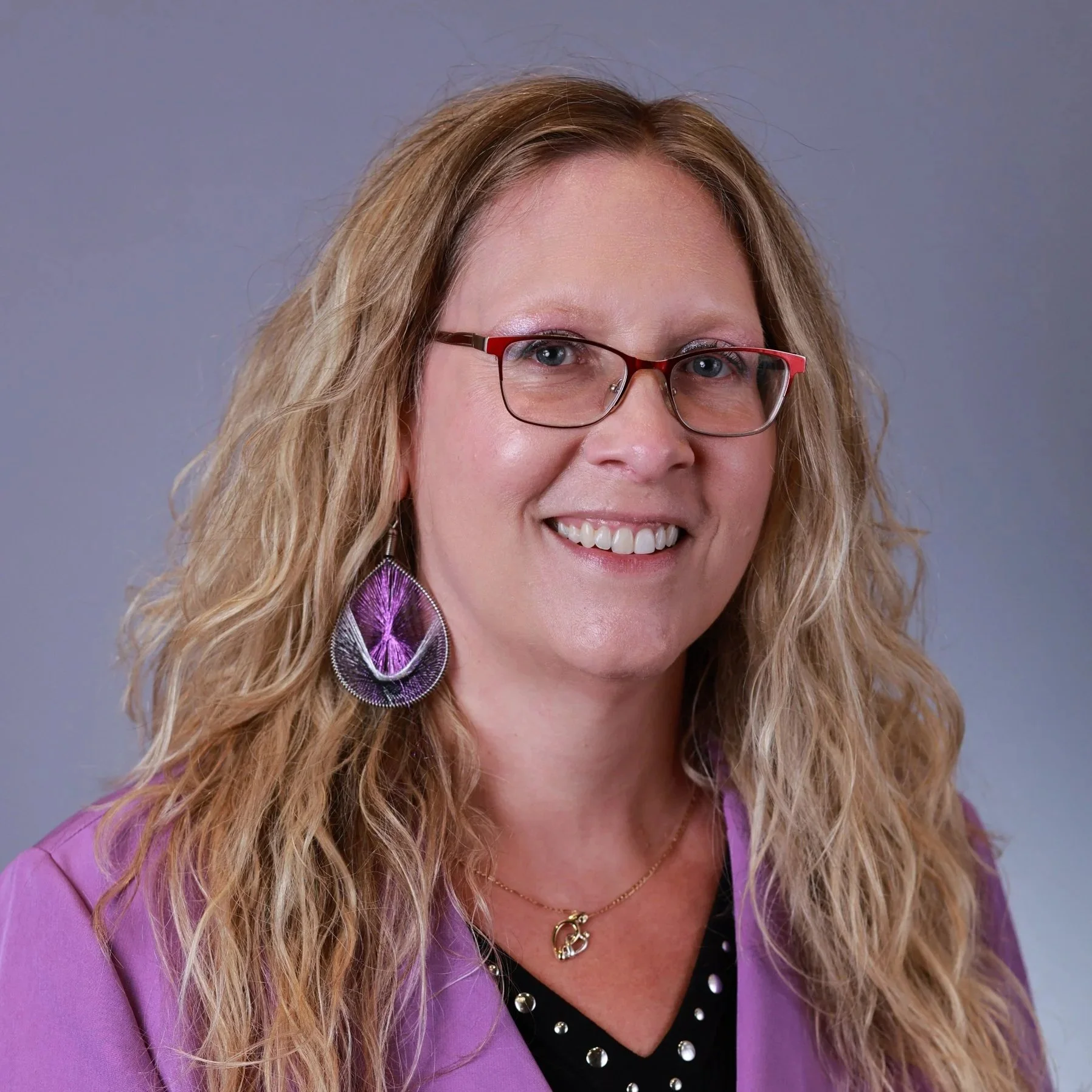 A woman with curly blonde hair, wearing glasses, smiling, and wearing a necklace, against a sparkly dark background.