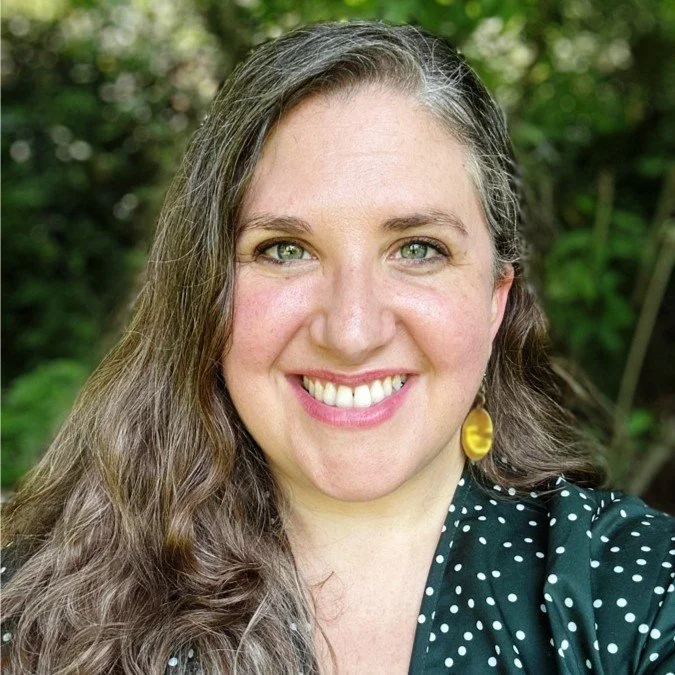 A woman with curly brown hair and blue eyes smiling at the camera, wearing a dark top, in front of a wooden wall.
