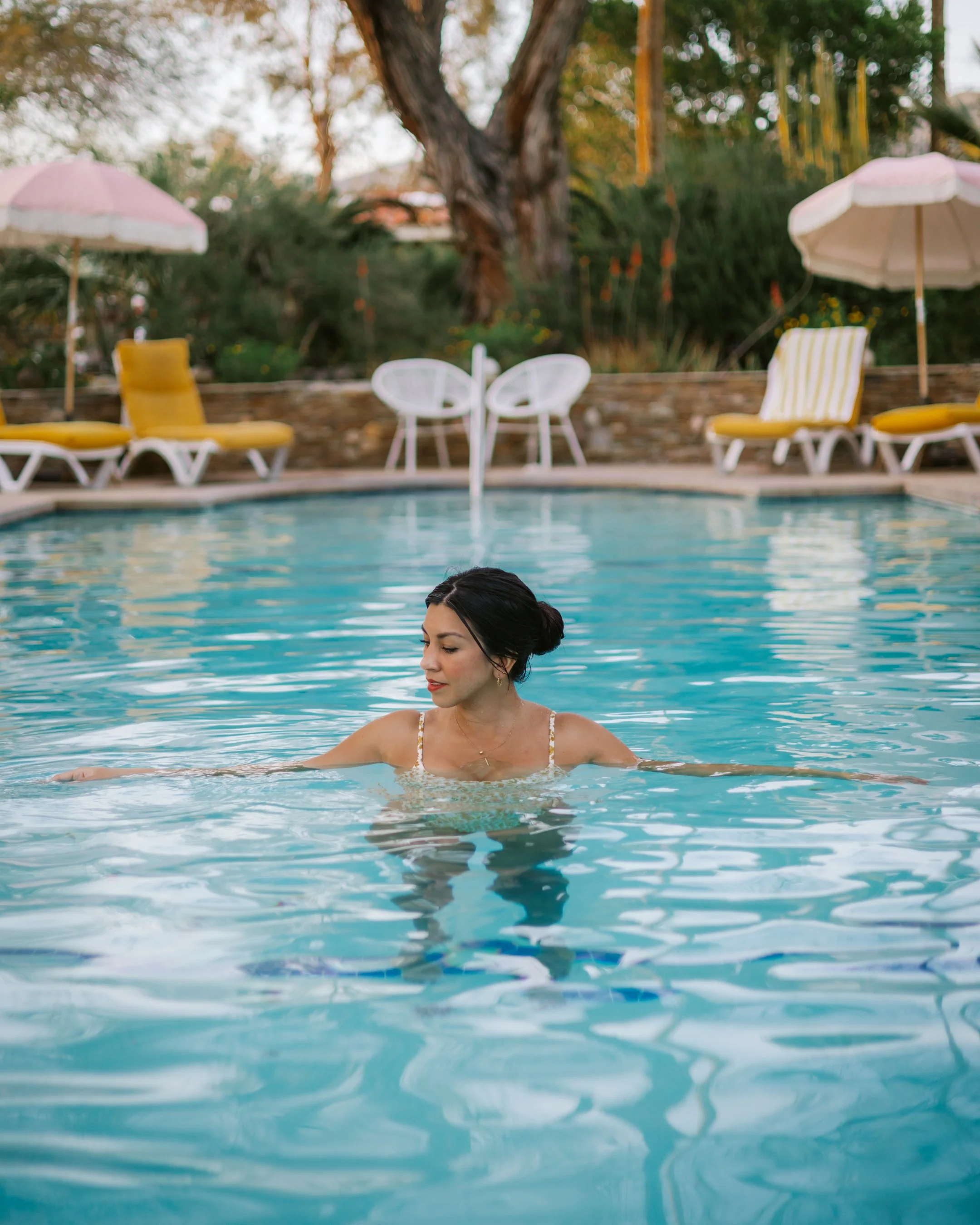 A woman with dark hair in a bun is swimming in a pool. In the background, there are yellow lounge chairs with striped cushions, white umbrellas, and lush green trees.