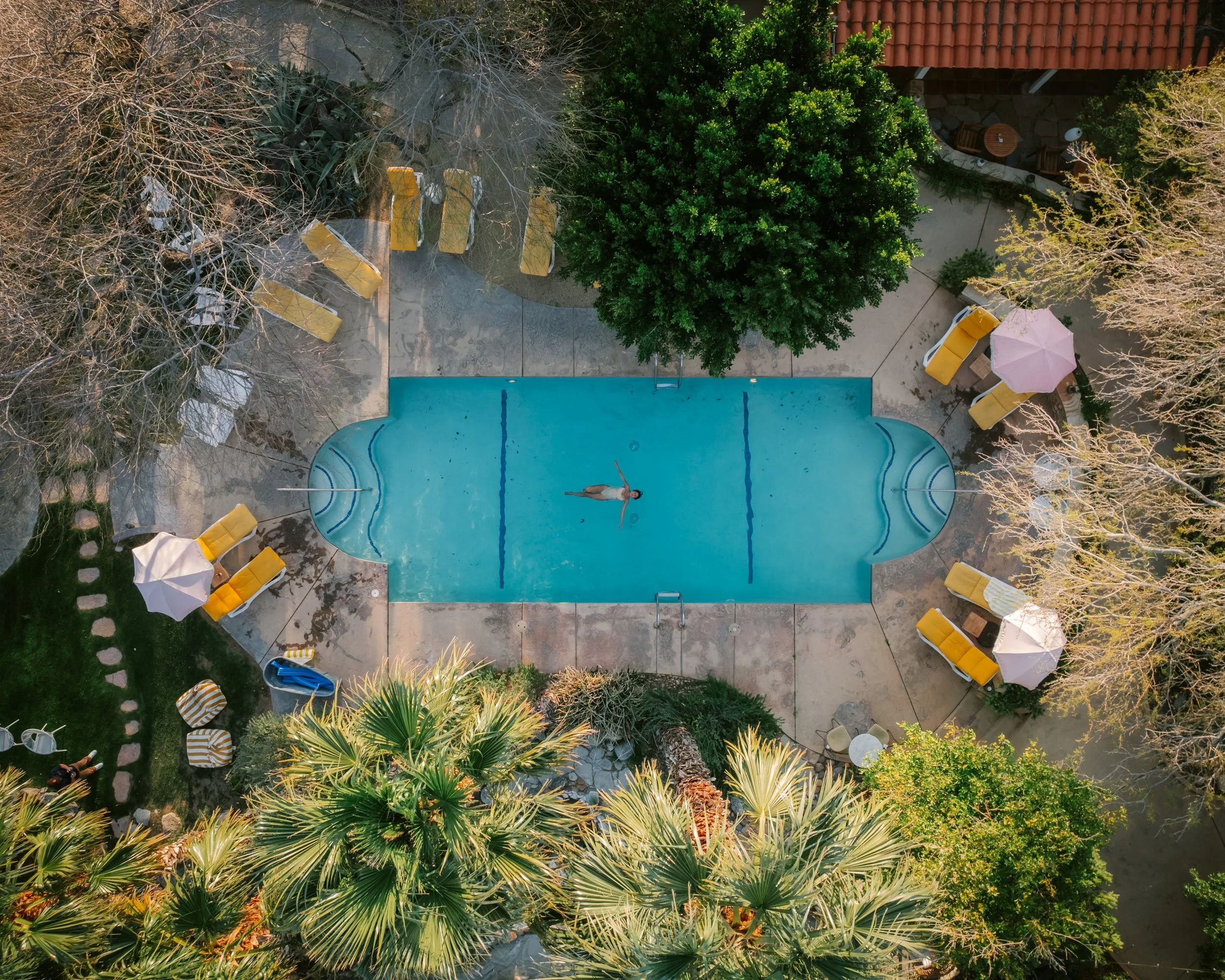 An aerial view of a backyard swimming pool with a person floating on their back, surrounded by outdoor furniture, umbrellas, and trees.