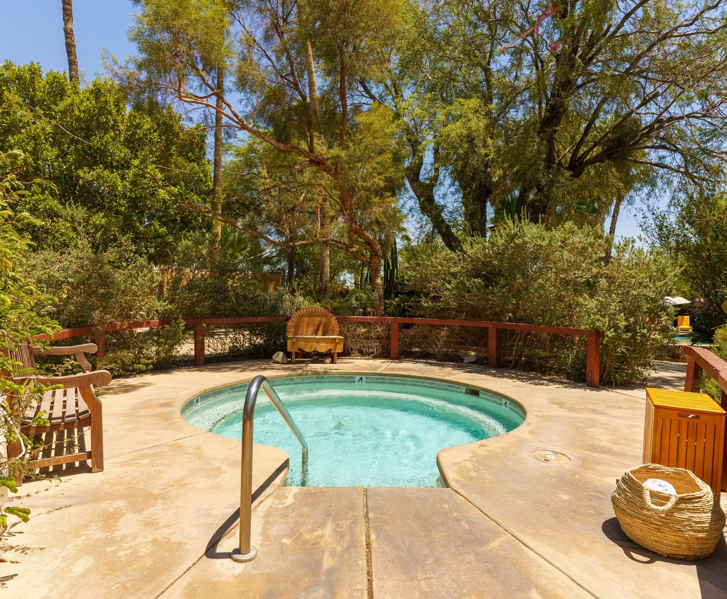 A backyard scene with a small round swimming pool, wooden deck chairs, a wicker basket, lush green trees, and a clear blue sky.