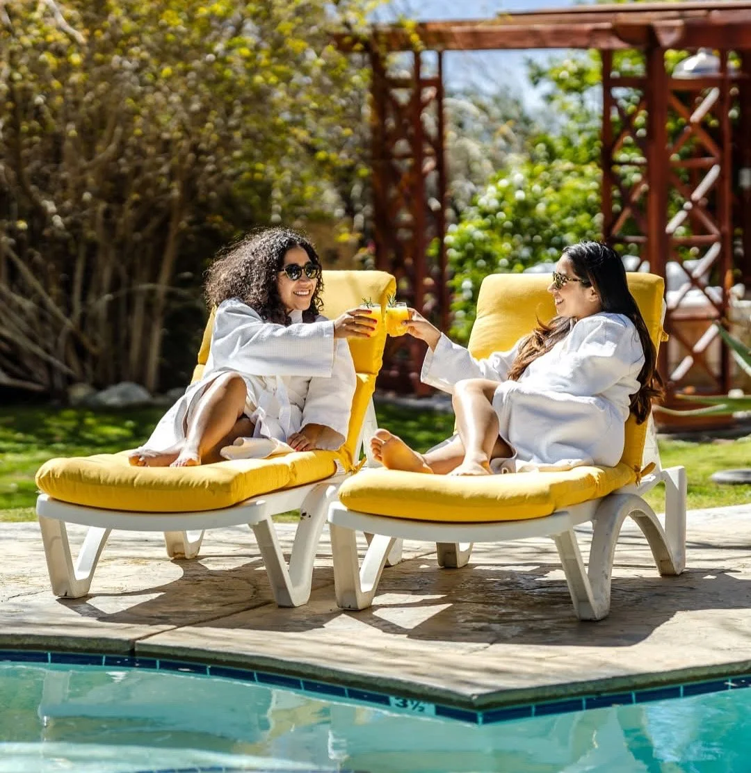Two women in white robes relaxing on yellow lounge chairs by a pool, enjoying drinks and smiling at each other on a sunny day with lush greenery in the background.