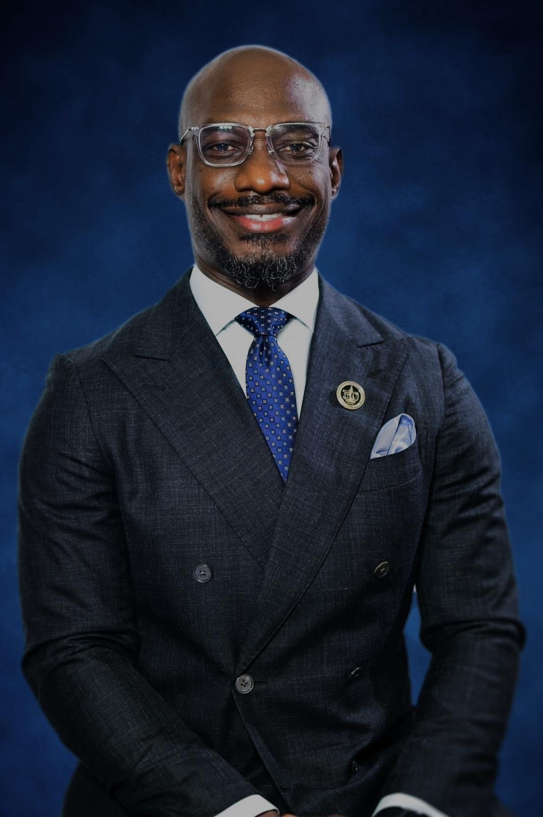 A professionally dressed man with glasses smiling in front of a dark blue background.