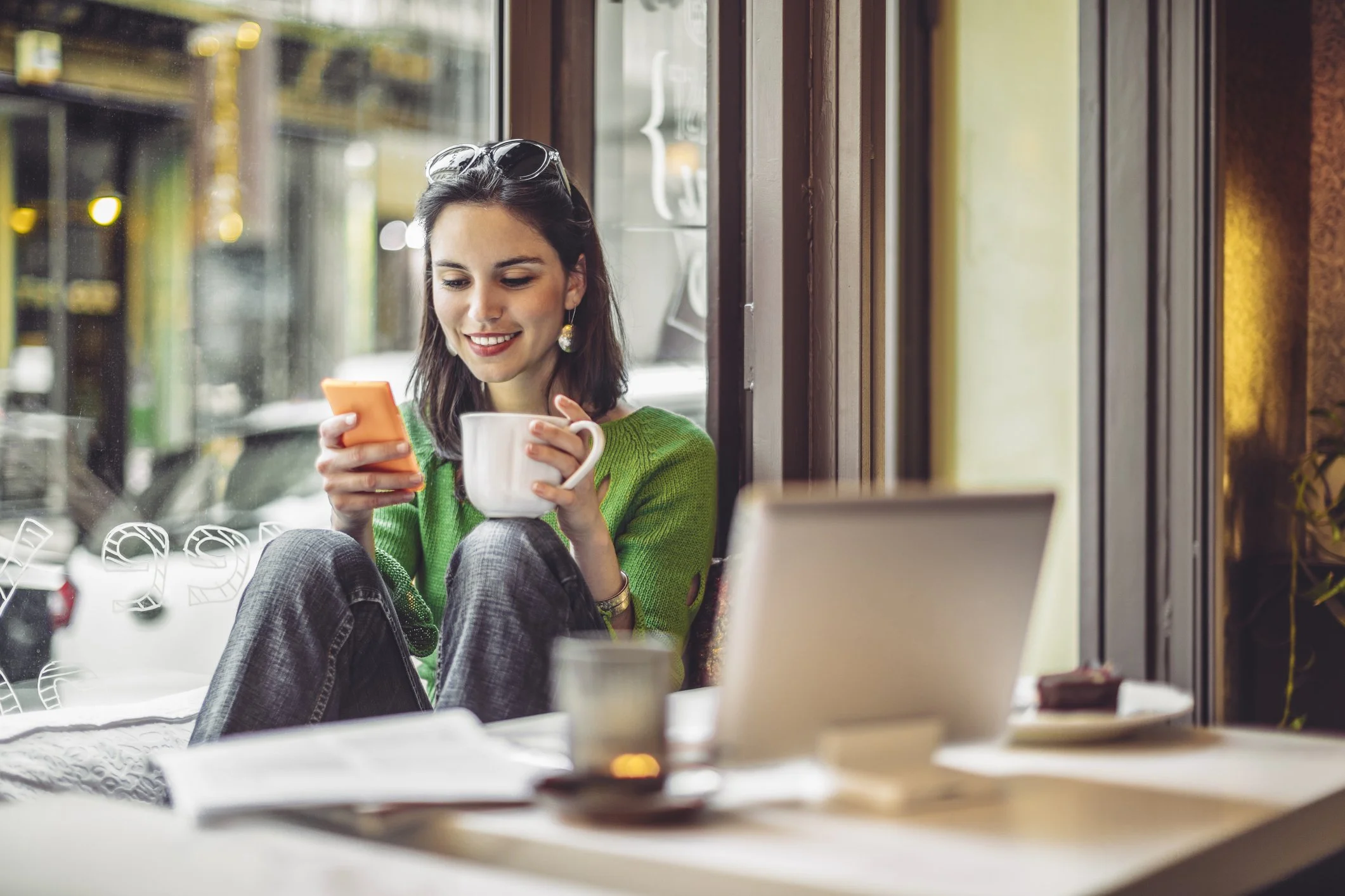 Woman in green sweater at a coffee shop ignoring her laptop and instead on her phone with a coffee in her hand.