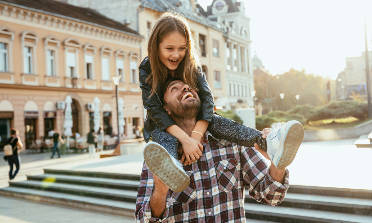 Middle-aged father with young daughter on his shoulders walking through historic European city centre during golden hour.