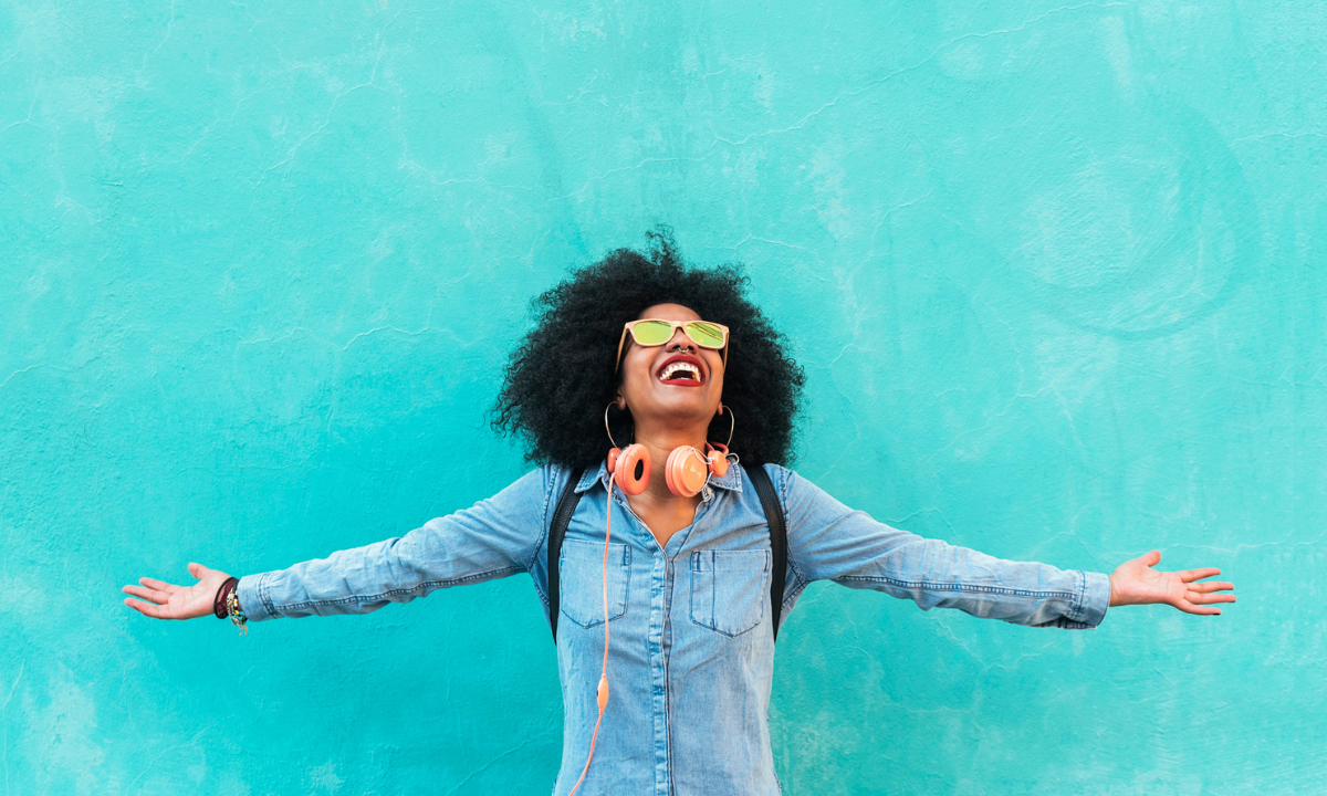 Young woman in a jean shirt and orange headphones with arms stretched, smiling at the sky.