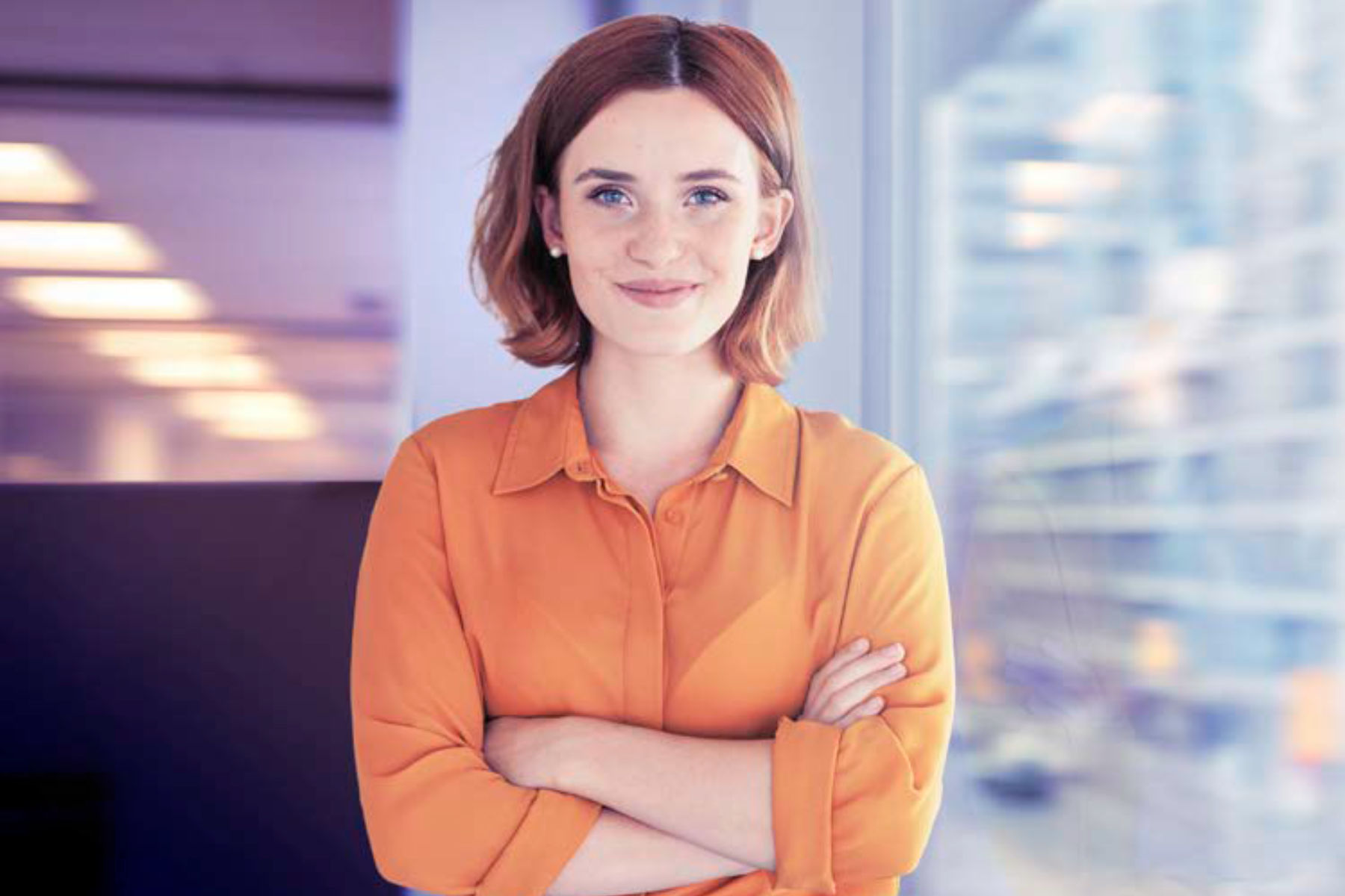 Red-haired woman in orange shirt with arms crossed staring at the camera with a smile.