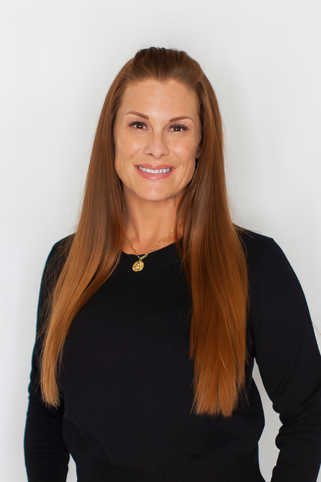 A woman with long red hair wearing a black top and a gold necklace smiling at the camera