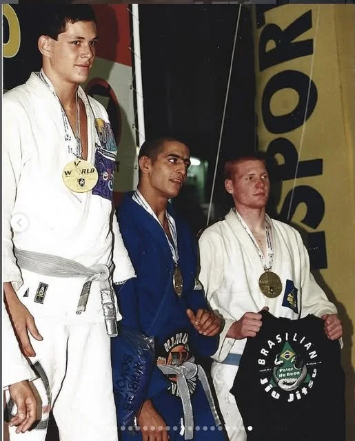Three male judo athletes on the podium with medals, wearing judo gi uniforms, one holding a black martial arts jacket, at a Brazilian jiu-jitsu competition.