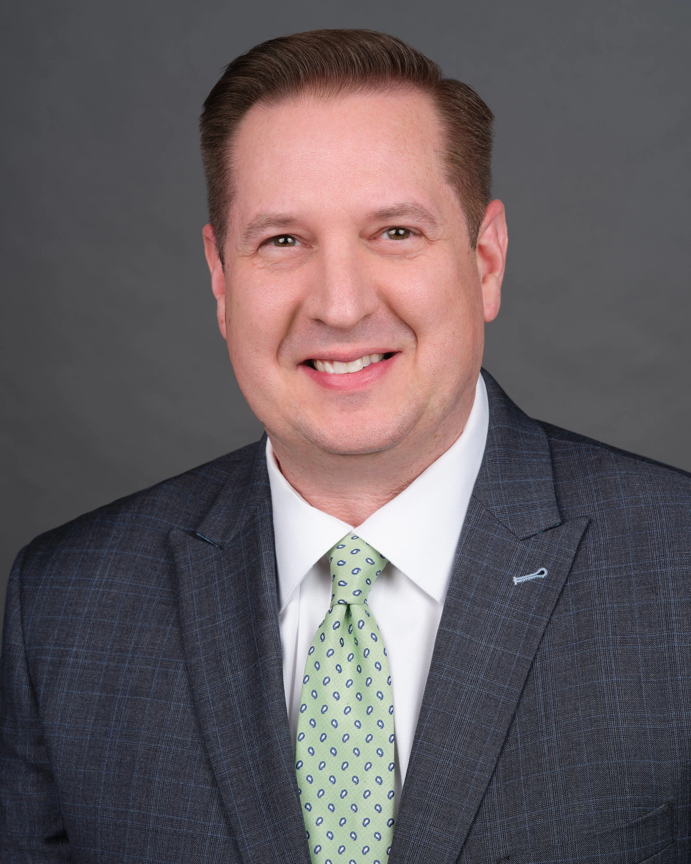 A headshot of a man with short, neatly combed brown hair, smiling, wearing a dark checkered suit, white shirt, and a light green tie with dark and light blue dots, posed against a plain dark background.
