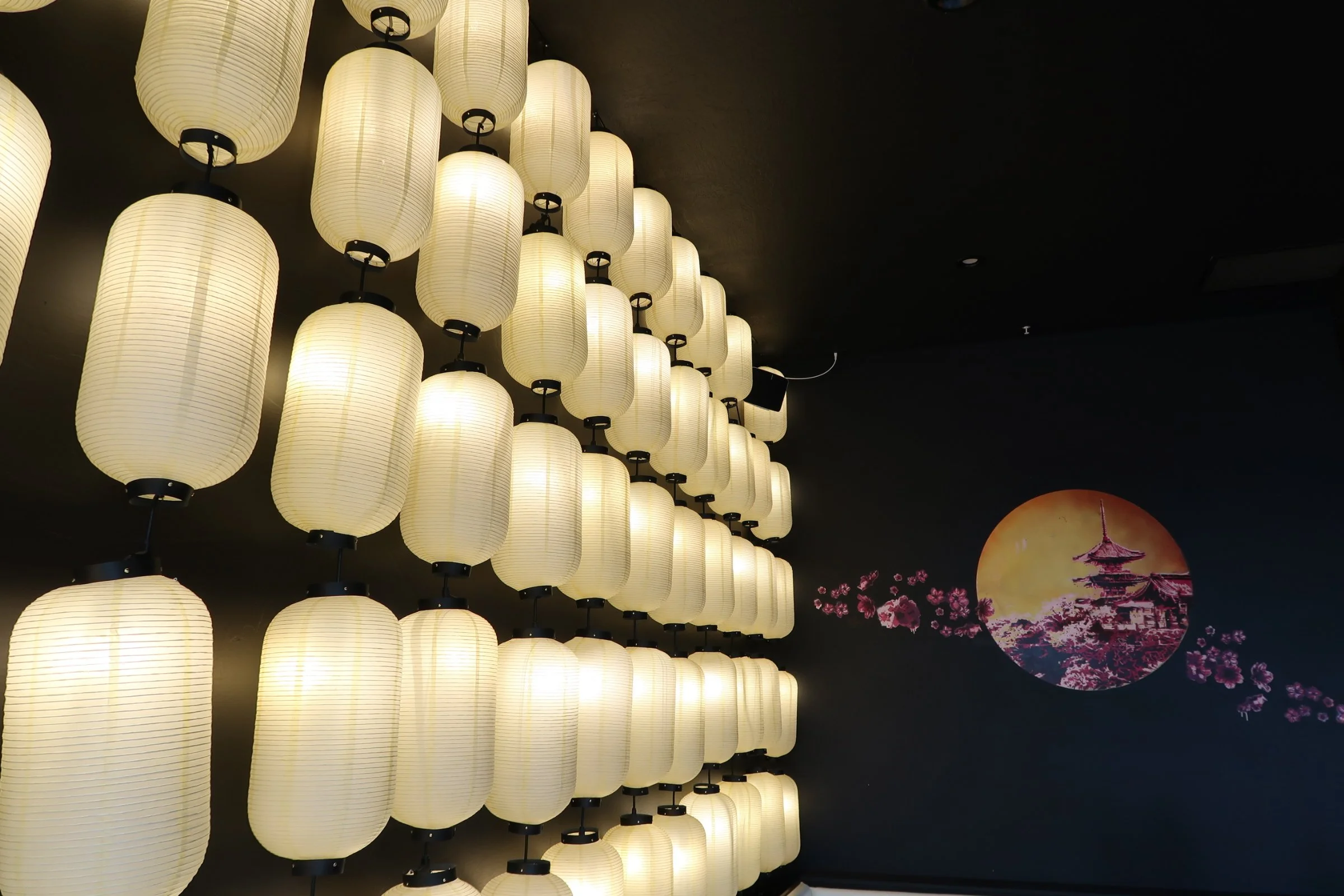 Wall of illuminated Japanese paper lanterns with a mural of a temple and cherry blossoms on a dark background.