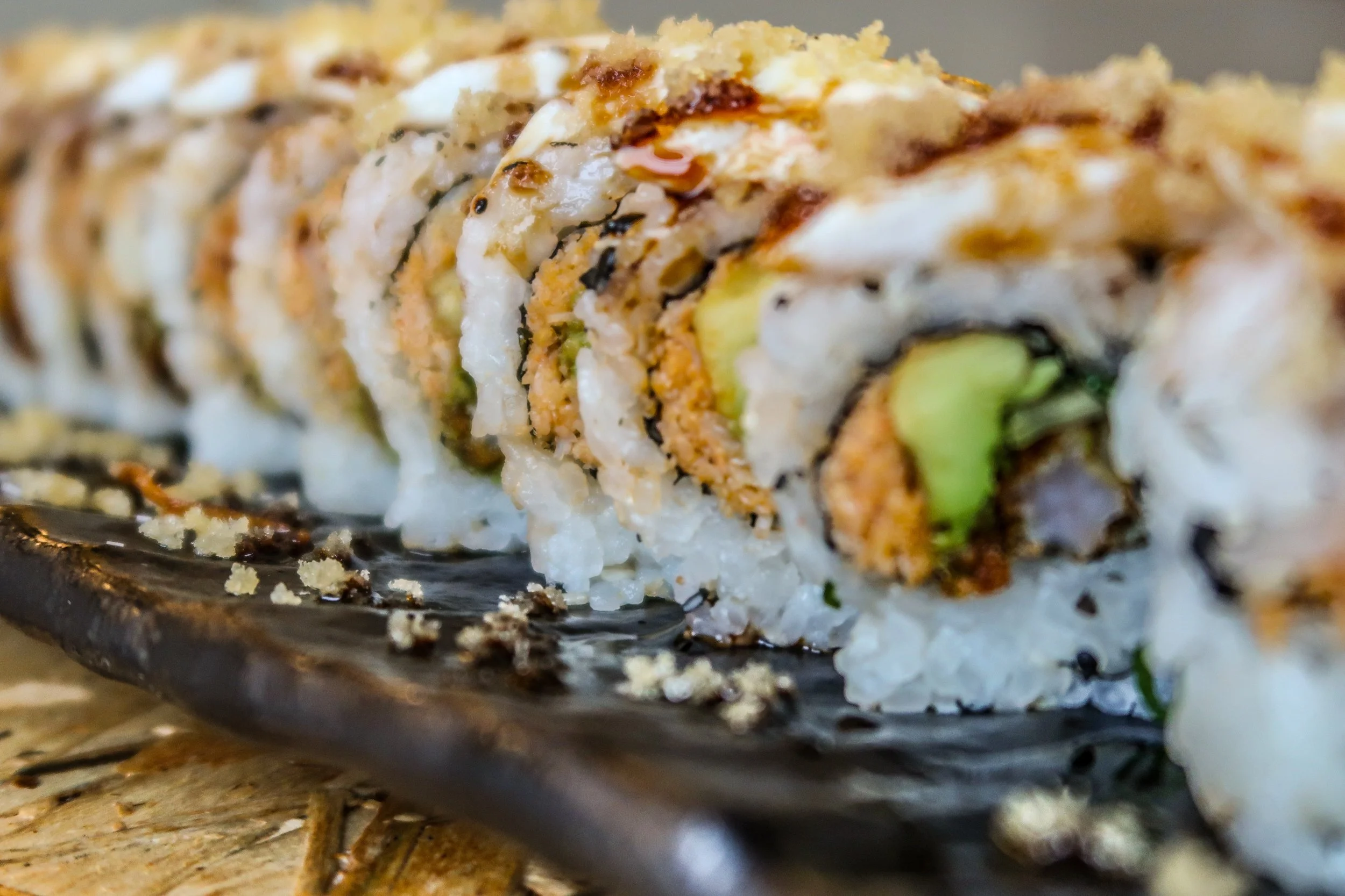 Close-up view of sushi roll with rice, avocado, crispy topping, and sauce on a black plate.